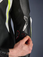 Tuta in pelle GP Tech V5 1PC, vista dettagliata della porta di connessione Tech-Air sulla manica, mano che mostra l'integrazione elettronica, pelle nera con pannelli traforati bianchi e giallo fluo, abbigliamento professionale da gara