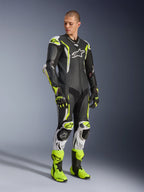Alpinestars GP Tech V5 One Piece Leather Suit, tuta in pelle intera, nero, bianco e giallo fluo, indossata da un modello con guanti e stivali da corsa coordinati, equipaggiamento professionale per gare motociclistiche con prestazioni aerodinamiche e protezioni.