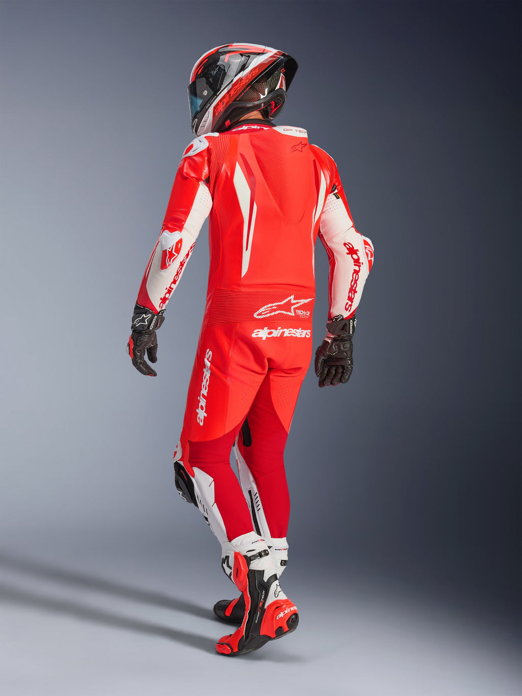 Tuta in pelle intera Alpinestars GP Tech V5, rosso fluo, rosso brillante e bianco, vista posteriore di un motociclista in equipaggiamento da gara completo, con gobba aerodinamica e pannelli bianchi su maniche e gambe, abbinata a un casco coordinato e stivali da corsa rossi, design professionale per prestazioni in pista.