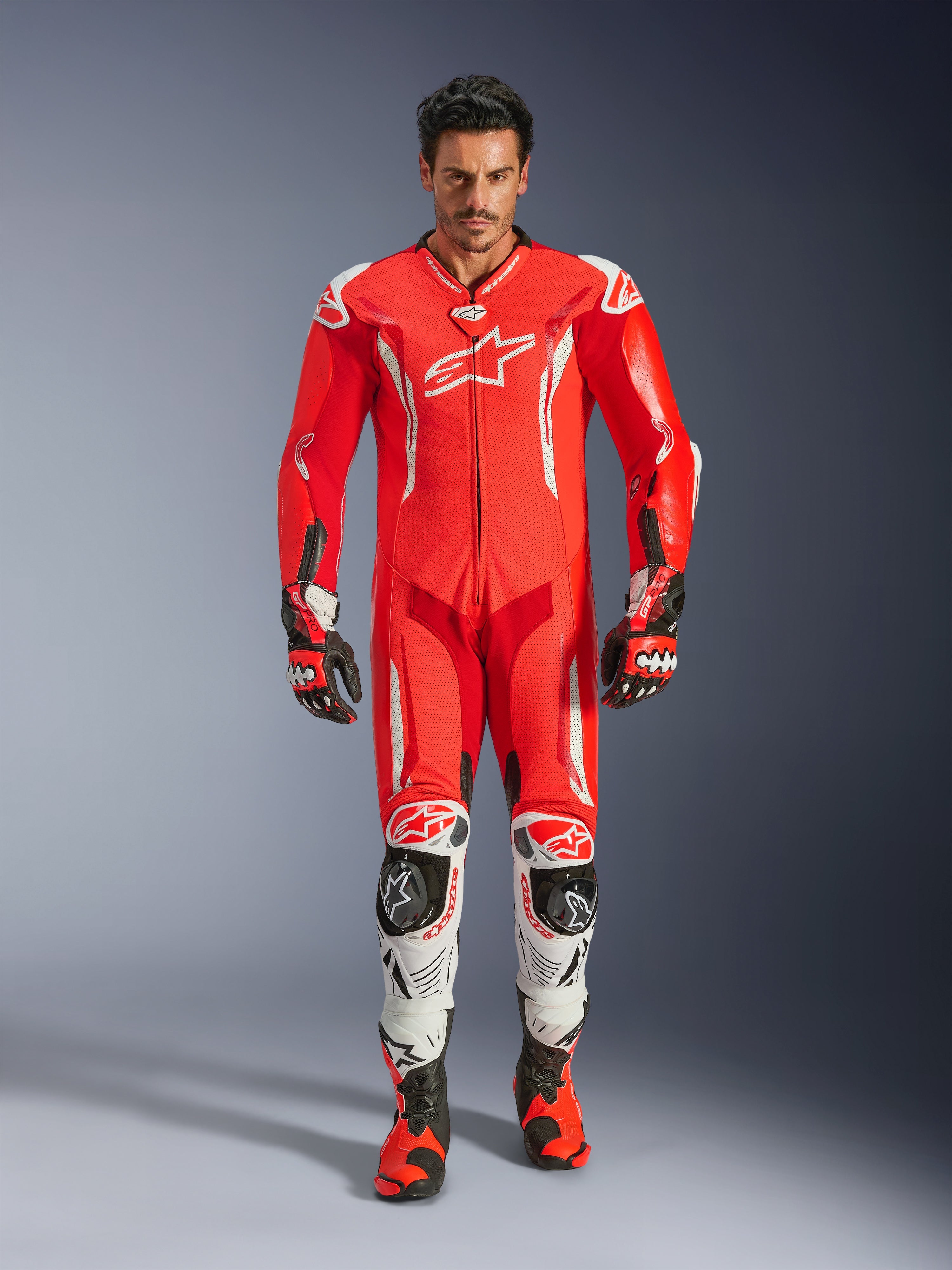 Alpinestars GP Tech V5, tuta professionale da corsa in pelle monopezzo, rosso fluo e rosso brillante con accenti bianchi, indossata da un pilota con stivali e guanti coordinati, con gobba aerodinamica, costruzione in pelle premium e protezioni avanzate per prestazioni in pista
