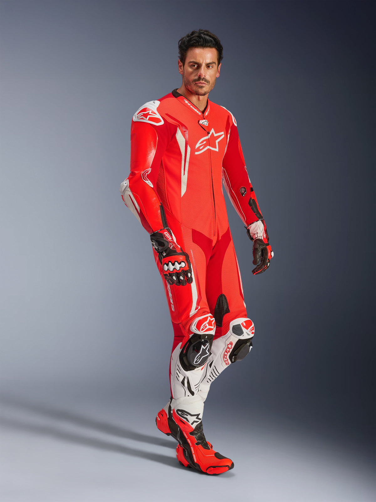 Uomo con tuta in pelle Alpinestars GP Tech V5 1PC, rosso fluo, rosso brillante e bianco, coordinata con guanti e stivali da corsa, abbigliamento professionale da moto con gobba aerodinamica e protezioni integrate.