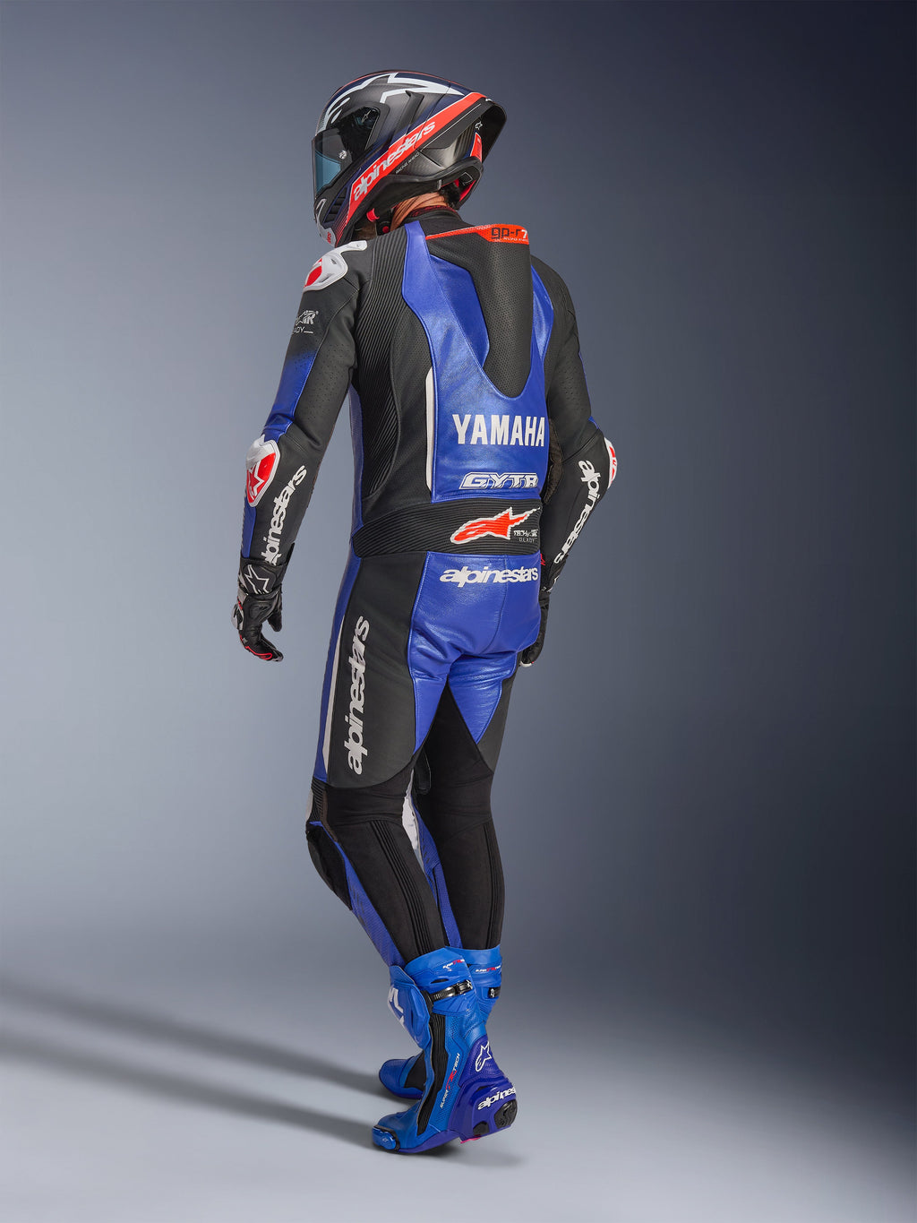 Tuta in pelle intera Alpinestars Yamaha GP-R7, blu metallizzato, nero e bianco, vista posteriore con gobba aerodinamica e marchio Yamaha, indossata con stivali e casco racing blu coordinati, tuta professionale in pelle per competizioni su pista