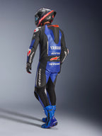 Tuta in pelle intera Alpinestars Yamaha GP-R7, blu metallizzato, nero e bianco, vista posteriore con gobba aerodinamica e marchio Yamaha, indossata con stivali e casco racing blu coordinati, tuta professionale in pelle per competizioni su pista