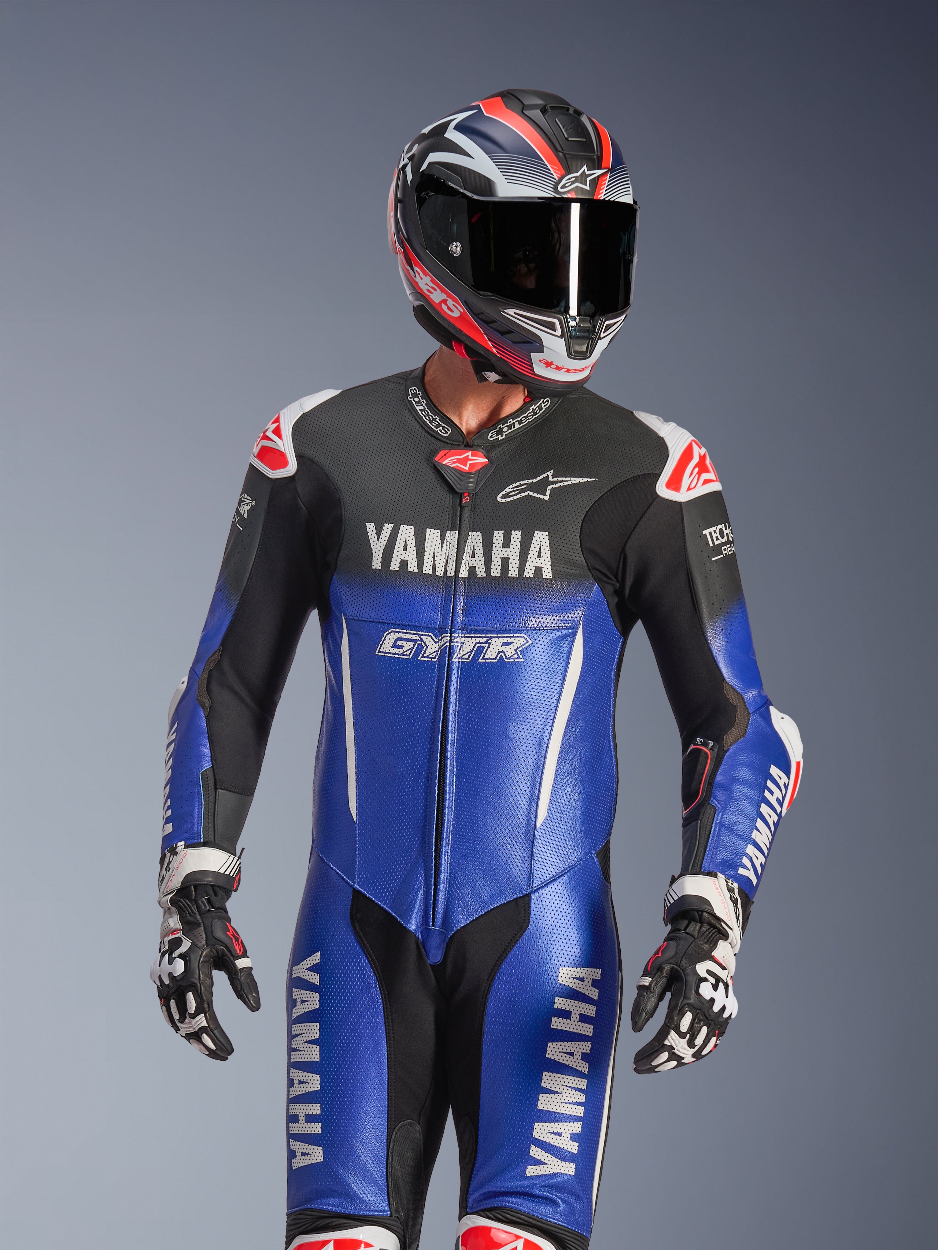 Tuta intera in pelle Yamaha GP-R7, blu metallizzato e nero con dettagli bianchi e rossi, indossata con casco e guanti da corsa coordinati, con marchi YAMAHA e GYTR, pannelli in pelle traforata e loghi Alpinestars, abbigliamento professionale da competizione motociclistica.