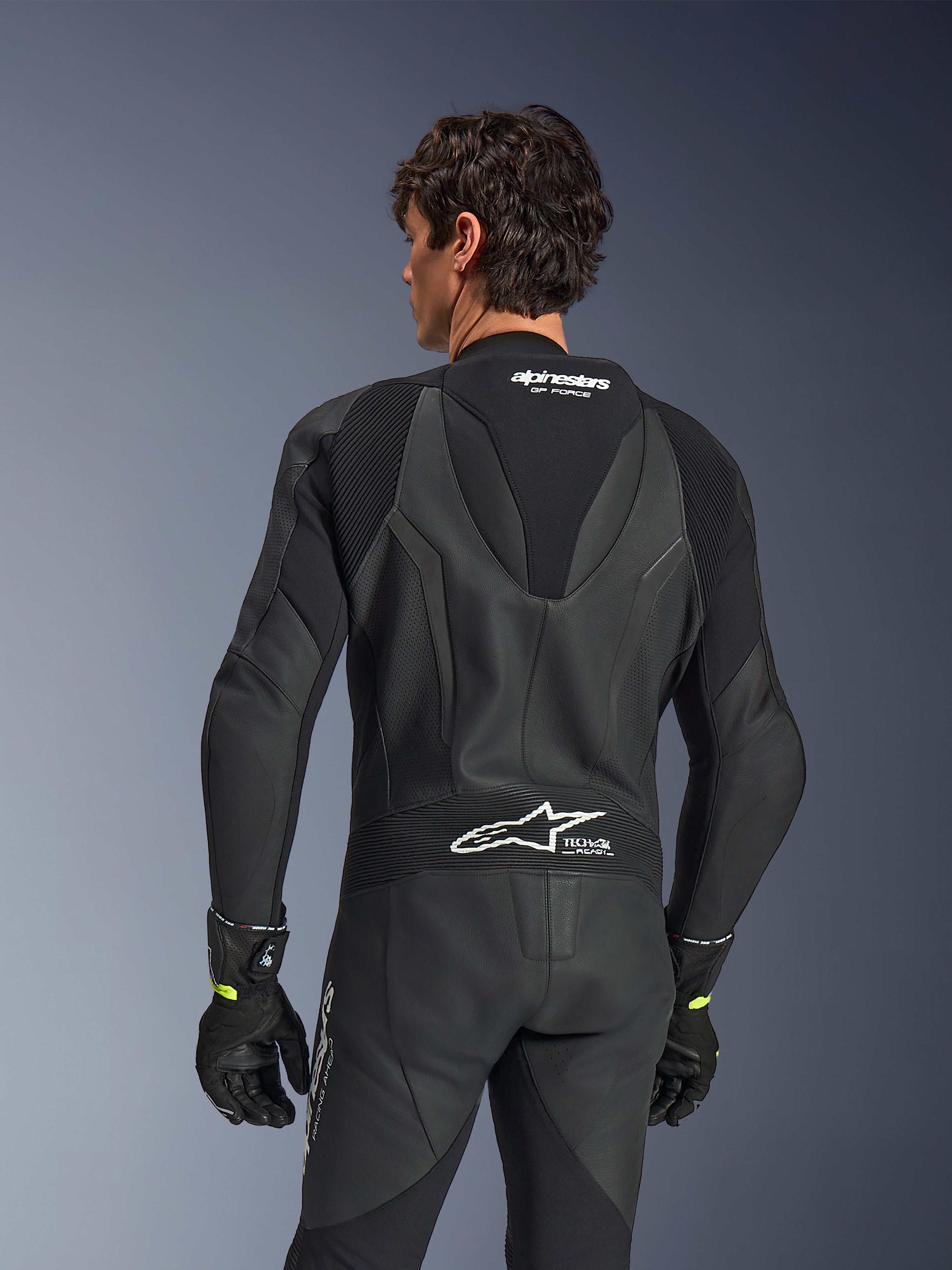 Tuta in pelle Alpinestars GP Force V2, intera, nera, vista posteriore con pelle traforata e pannelli elasticizzati a fisarmonica, loghi bianchi Alpinestars e Tech-Air Ready sulla parte superiore e inferiore della schiena, indossata con guanti neri, abbigliamento da competizione motociclistica ad alte prestazioni