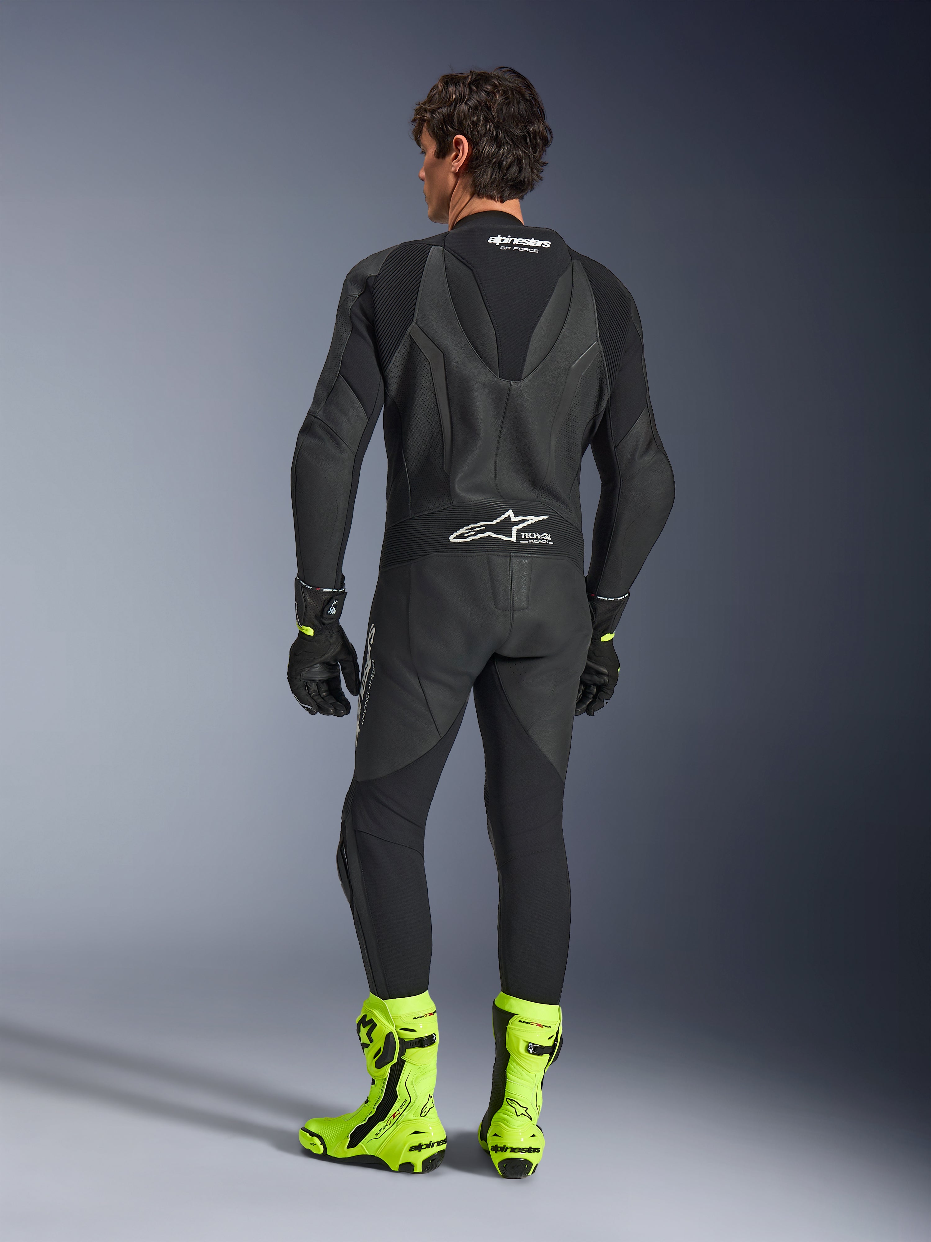 Alpinestars GP Force V2 Leather Suit - 1 pezzo, nero, vista posteriore indossata da un modello, con gobba aerodinamica e marchio Tech-Air Ready, abbinata a guanti neri e stivali da corsa giallo fluorescente, equipaggiamento professionale da pista.
