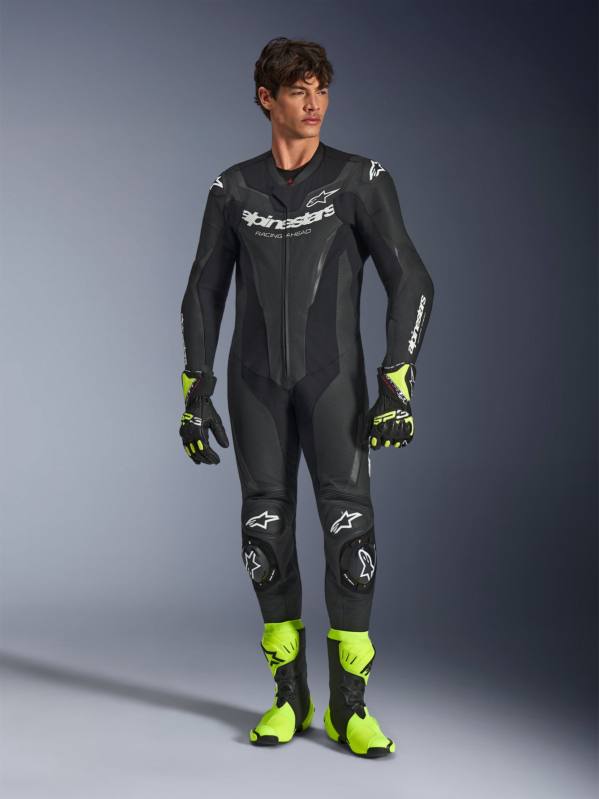 Alpinestars GP Force V2 Leather Suit, tuta da moto monopezzo in pelle nera, indossata con stivali e guanti giallo fluo, caratterizzata da loghi bianchi e pelle traforata, design aderente per le giornate in pista