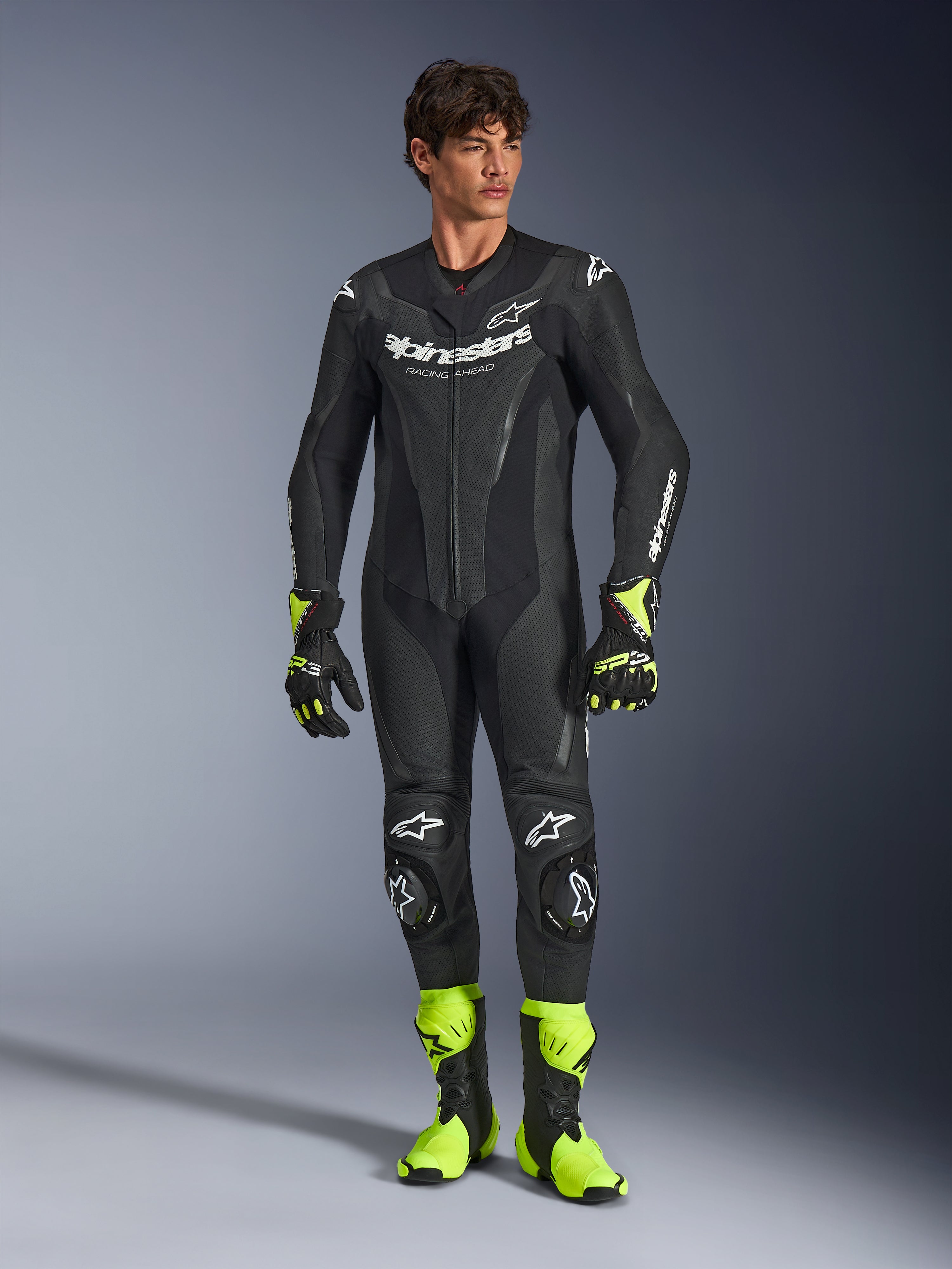 Alpinestars GP Force V2 Leather Suit, tuta da moto monopezzo in pelle nera, indossata con stivali e guanti giallo fluo, caratterizzata da loghi bianchi e pelle traforata, design aderente per le giornate in pista