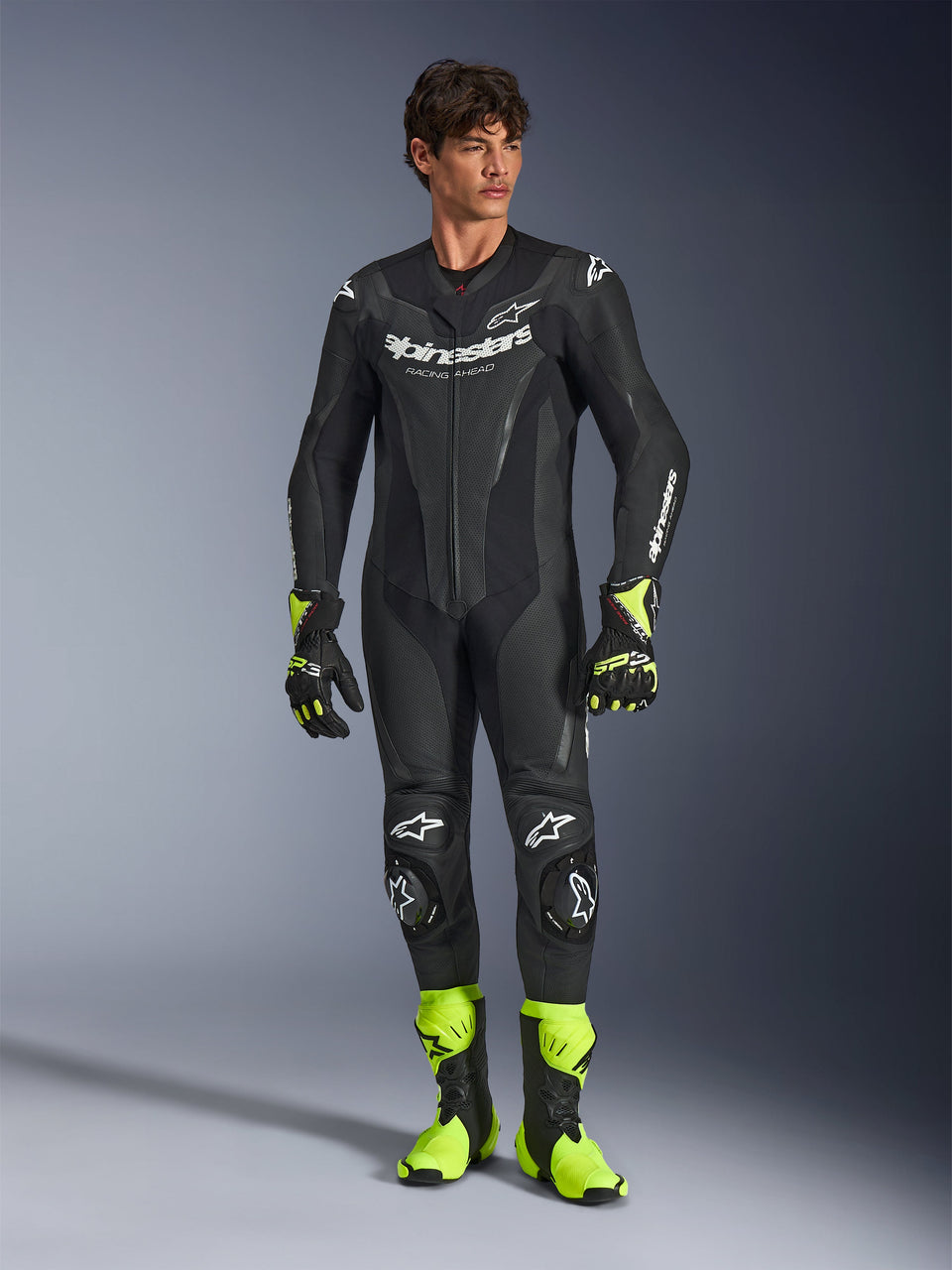 Alpinestars GP Force V2 Leather Suit, tuta da moto monopezzo in pelle nera, indossata con stivali e guanti giallo fluo, caratterizzata da loghi bianchi e pelle traforata, design aderente per le giornate in pista