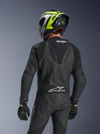 Alpinestars GP Force V2 Leather Suit 1-Piece, nero, vista posteriore che mostra la gobba aerodinamica e la struttura in pelle traforata, indossata con casco da corsa nero e giallo neon e guanti neri, dotata di pannelli elasticizzati a fisarmonica e logo Tech-Air Ready bianco.