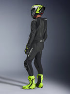 Pilota di moto con tuta in pelle Alpinestars GP Force V2 1-Piece in nero, vista posteriore laterale con loghi bianchi su coscia e parte bassa della schiena, gobba aerodinamica, abbinata a casco nero e giallo neon, guanti e stivali racing coordinati, abbigliamento professionale da pista e guida sportiva.