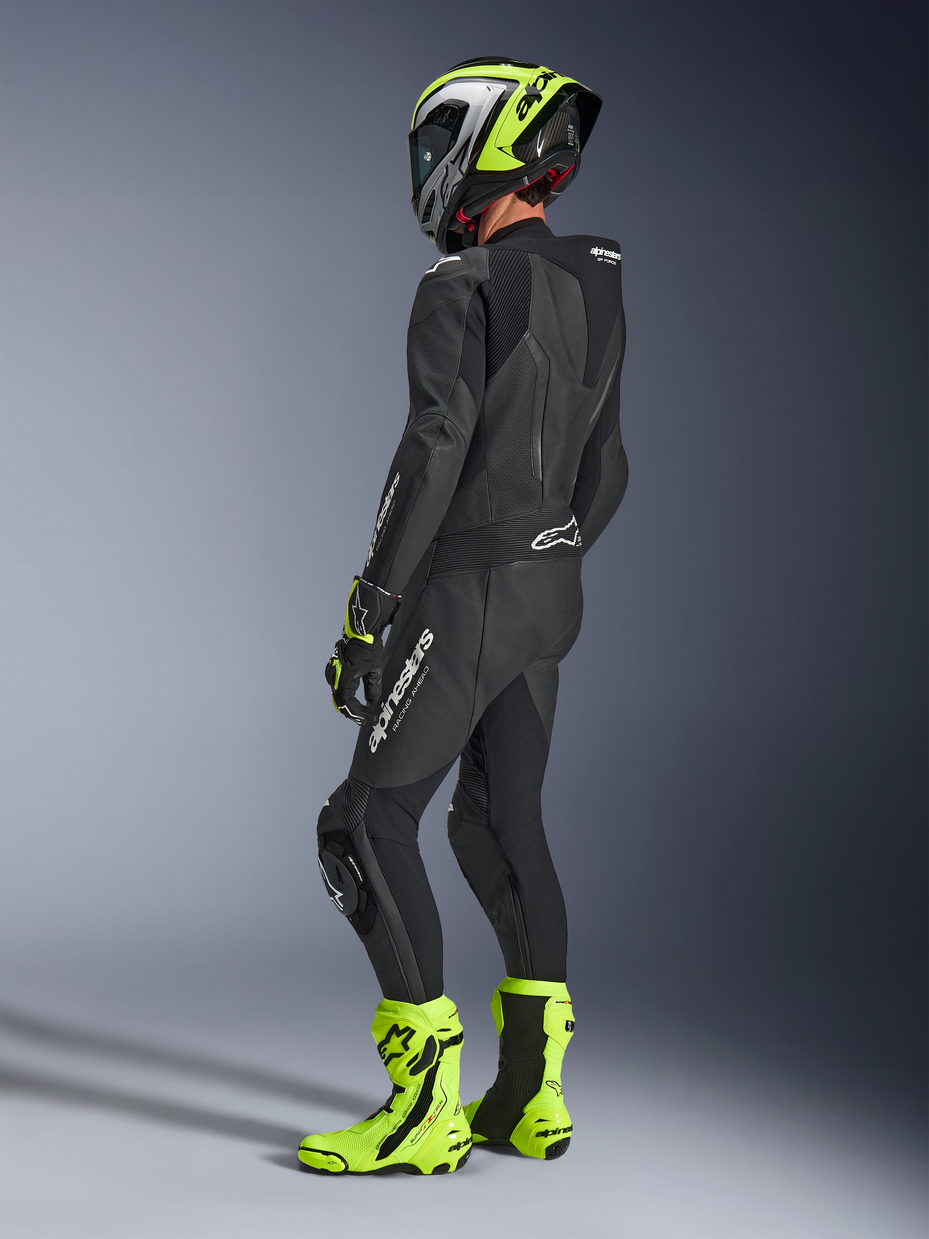 Pilota di moto con tuta in pelle Alpinestars GP Force V2 1-Piece in nero, vista posteriore laterale con loghi bianchi su coscia e parte bassa della schiena, gobba aerodinamica, abbinata a casco nero e giallo neon, guanti e stivali racing coordinati, abbigliamento professionale da pista e guida sportiva.