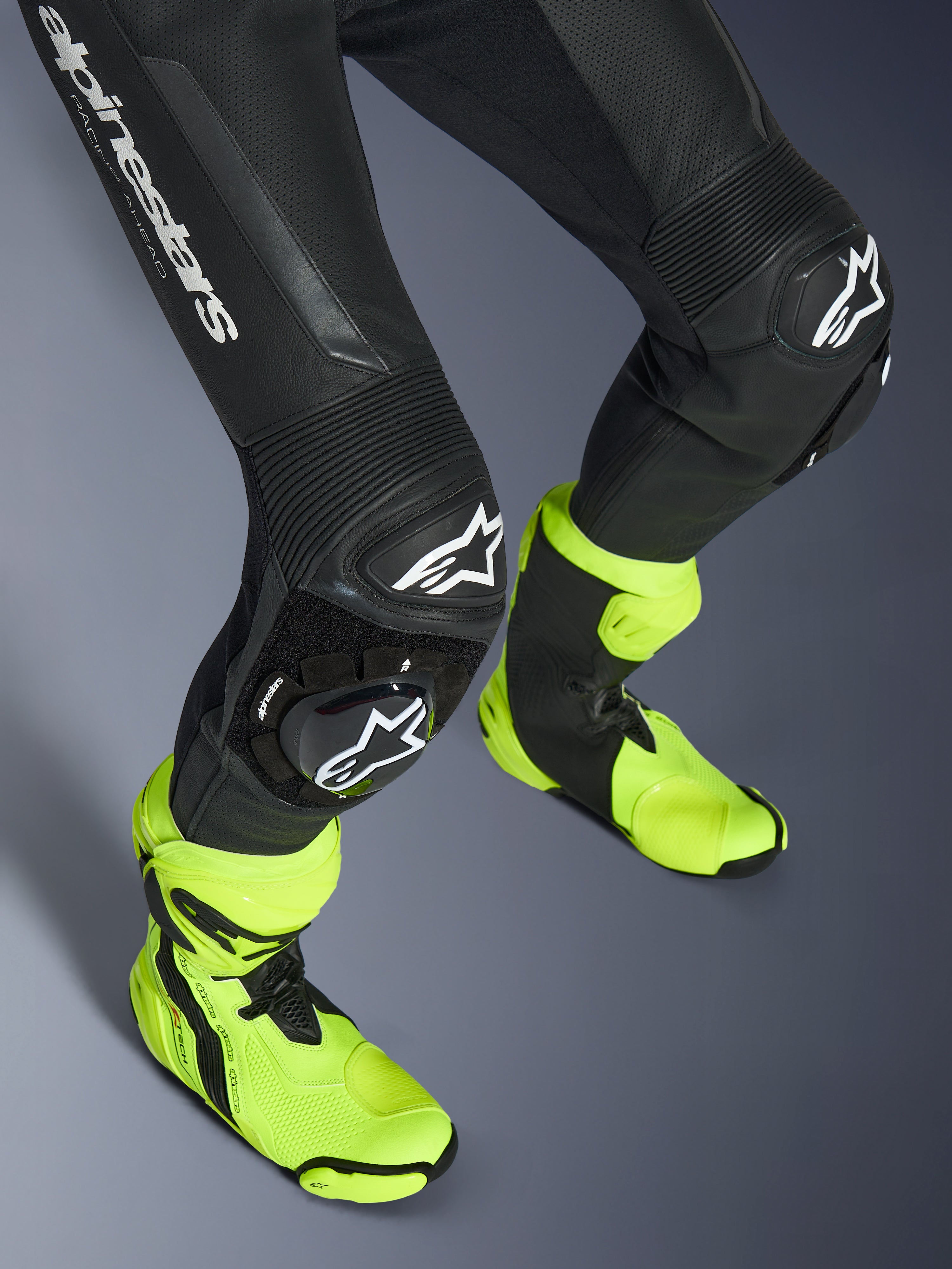 Tuta in pelle Alpinestars GP Force V2, tuta in pelle nera monopezzo, primo piano di gambe e ginocchia con saponette con logo bianco, pannelli elasticizzati a fisarmonica, indossata con stivali da corsa giallo neon brillante, equipaggiamento da corsa motociclistica ad alte prestazioni