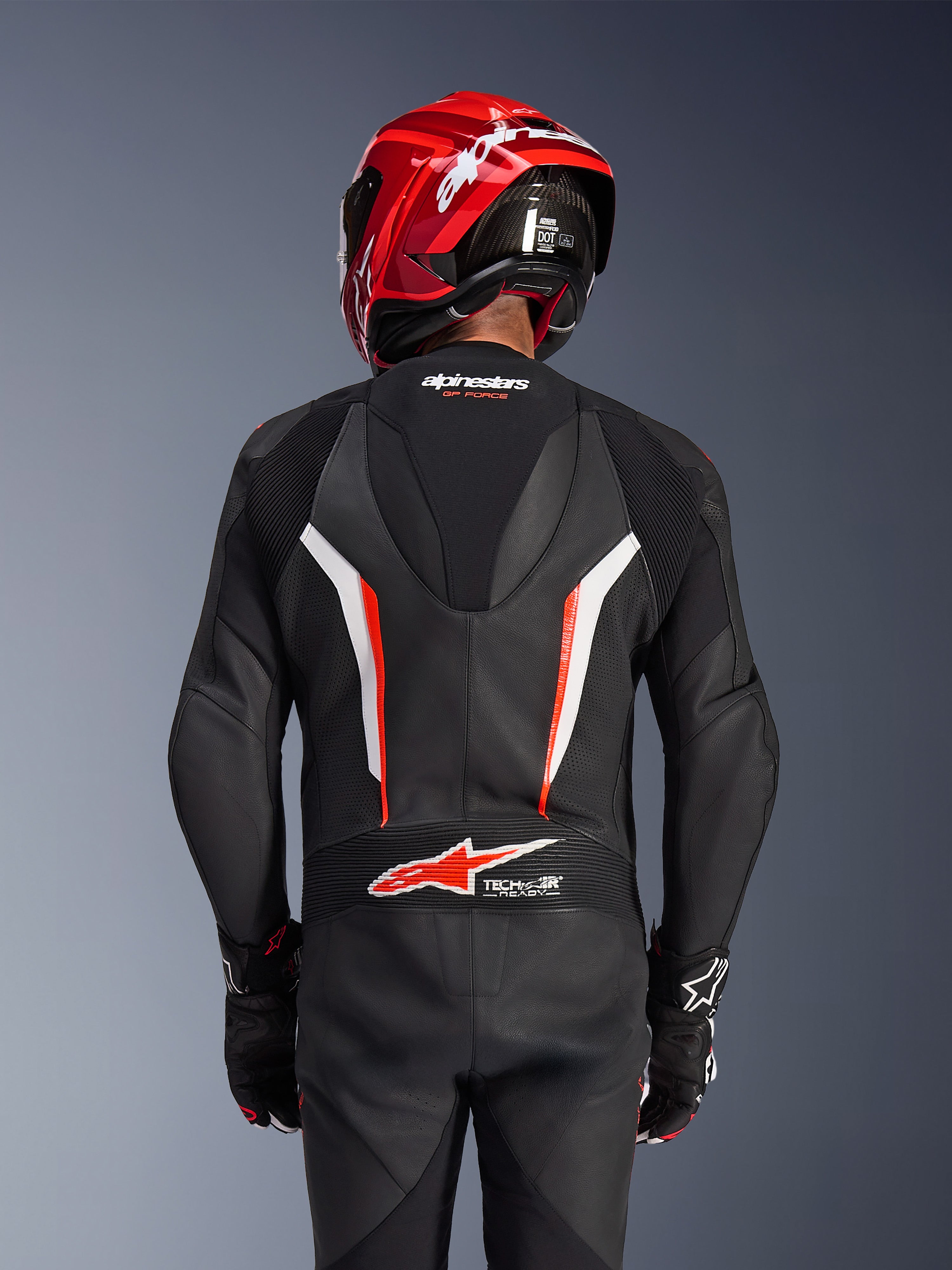 Tuta in pelle intera Alpinestars GP Force V2, nero, bianco e rosso fluo, vista posteriore del pilota con casco da corsa rosso e guanti, con pannelli in pelle traforata, gobba aerodinamica con marchio Alpinestars e logo Tech-Air Ready sulla parte bassa della schiena, abbigliamento professionale da gara motociclistica.