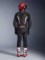 Pilota visto da dietro con tuta in pelle Alpinestars GP Force V2 1-Piece in nero, bianco e rosso fluo, caratterizzata da gobba aerodinamica e marchio Tech-Air Ready, coordinata con casco racing rosso e stivali bianchi e rossi, design professionale per attrezzatura da pista.