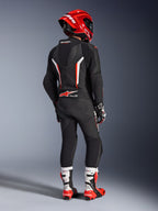 Pilota visto da dietro che indossa la tuta in pelle intera Alpinestars GP Force V2 in nero, bianco e rosso fluo, con gobba aerodinamica e pannelli in pelle traforata, abbinata a un casco Alpinestars rosso, guanti racing e stivali da moto rossi e bianchi, abbigliamento professionale da gara su pista.