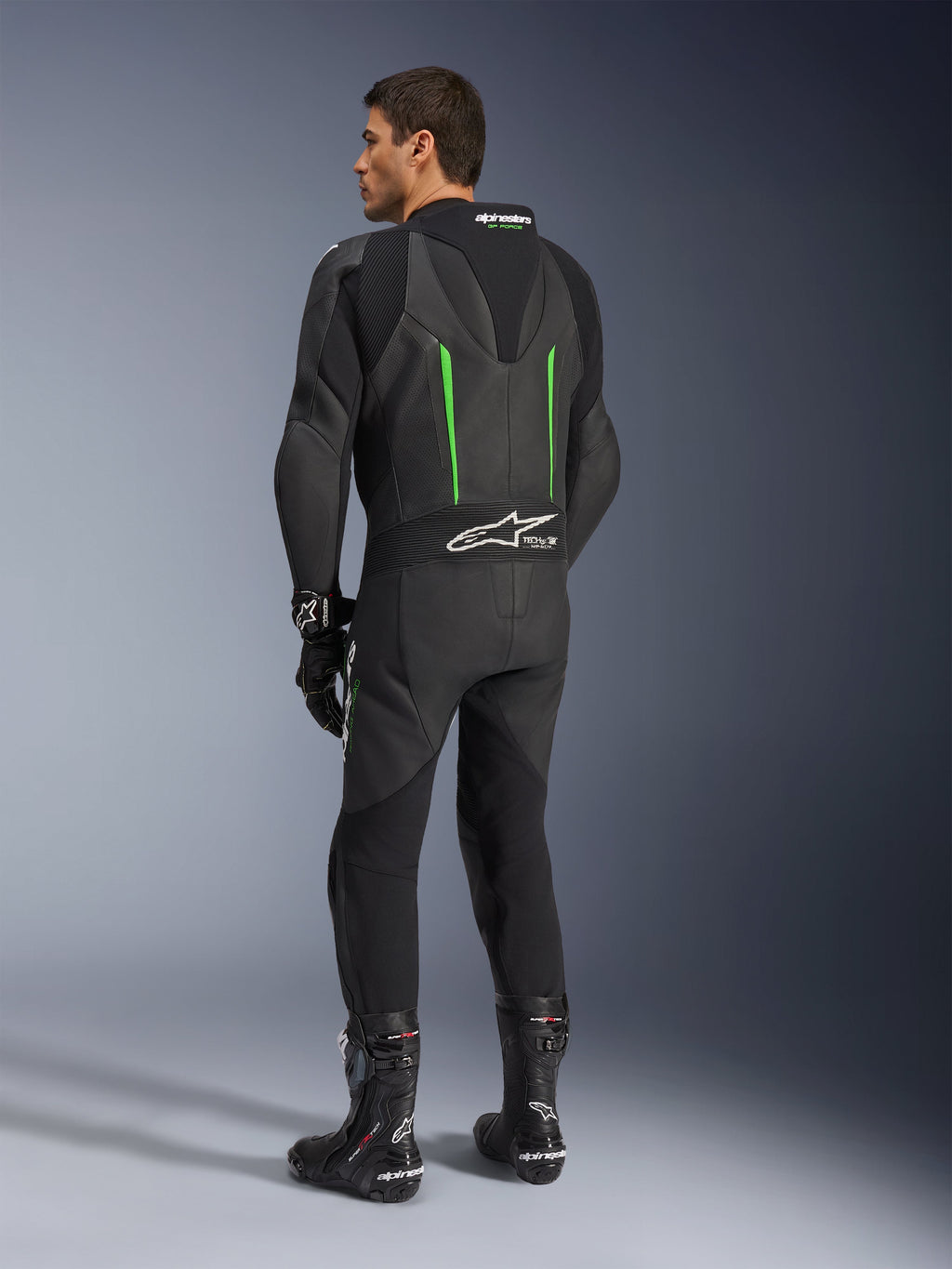 Tuta in pelle Alpinestars GP Force V2, tuta da moto intera, nero con dettagli verdi, vista posteriore che mostra la gobba aerodinamica e il logo bianco sul pannello elastico a fisarmonica, abbinata a stivali e guanti da corsa neri.