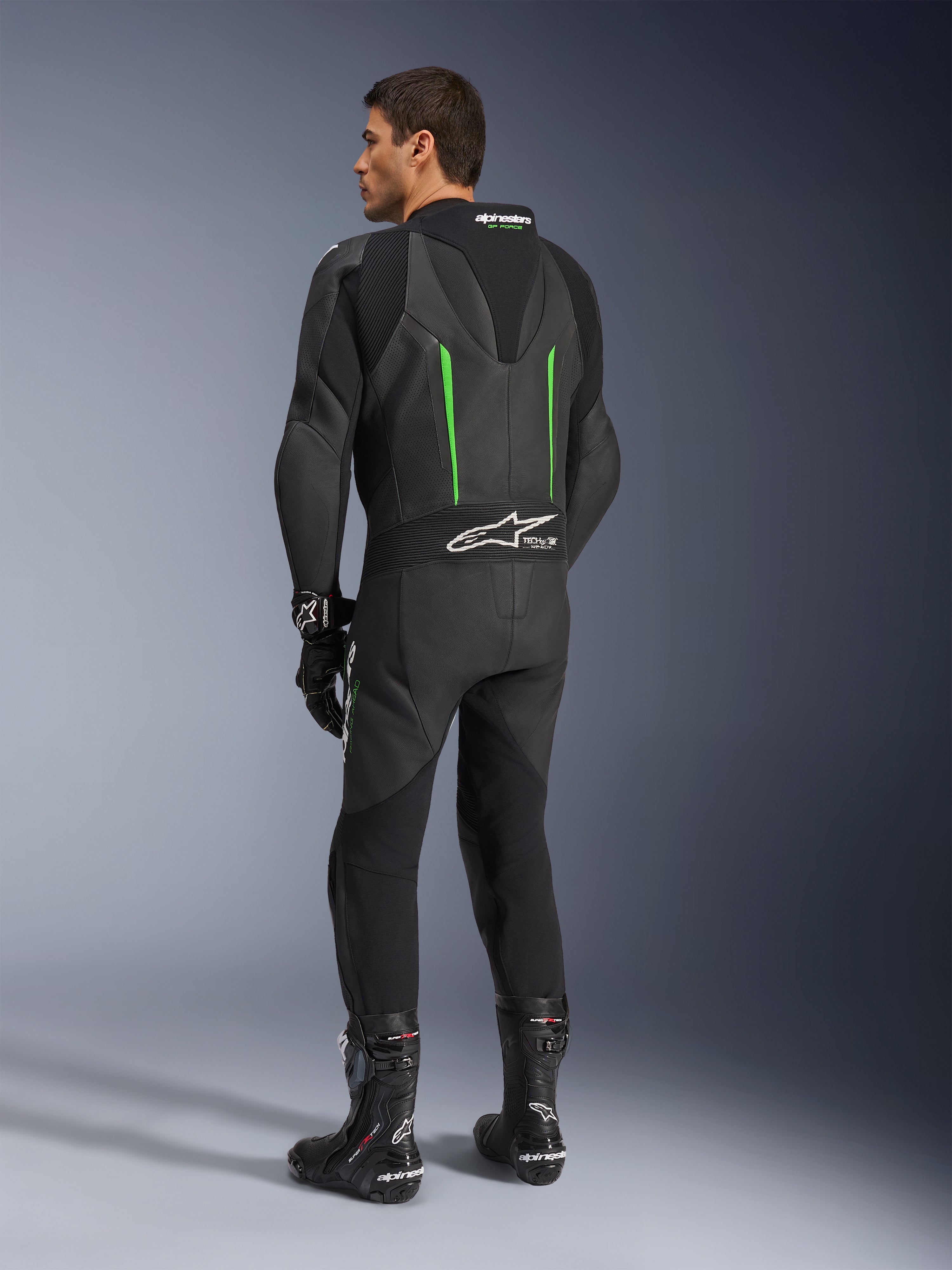 Tuta in pelle Alpinestars GP Force V2, tuta da moto intera, nero con dettagli verdi, vista posteriore che mostra la gobba aerodinamica e il logo bianco sul pannello elastico a fisarmonica, abbinata a stivali e guanti da corsa neri.