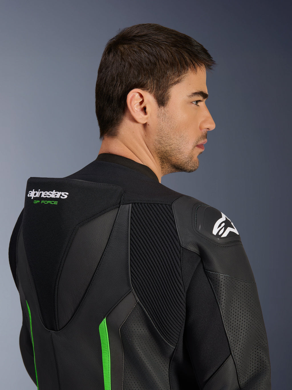 Primo piano della parte superiore della schiena e della spalla di un modello che indossa la tuta in pelle Alpinestars GP Force V2 in nero e verde, con gobba aerodinamica, marchio 'GP FORCE' verde, logo Astars bianco sulla spalla e pannelli a fisarmonica elasticizzati.