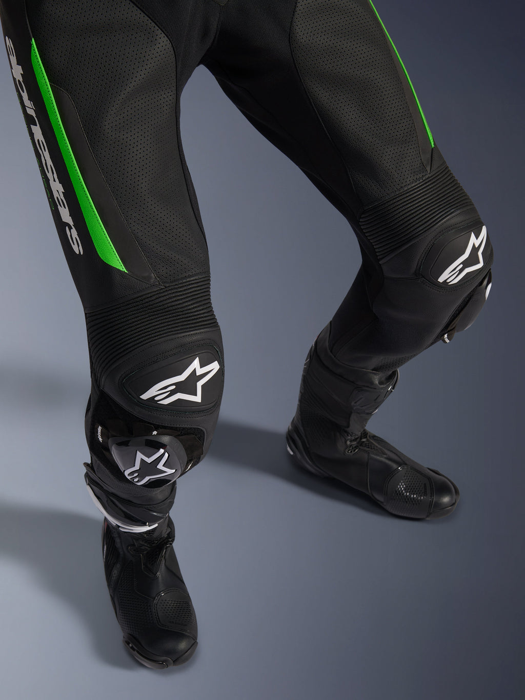 Alpinestars GP Force V2 Leather Suit - 1 Pezzo, primo piano delle gambe in nero e verde, con costruzione in pelle traforata, pannelli a fisarmonica elasticizzati sulle ginocchia, marchio Alpinestars bianco e saponette integrate, indossata con stivali da racing neri.