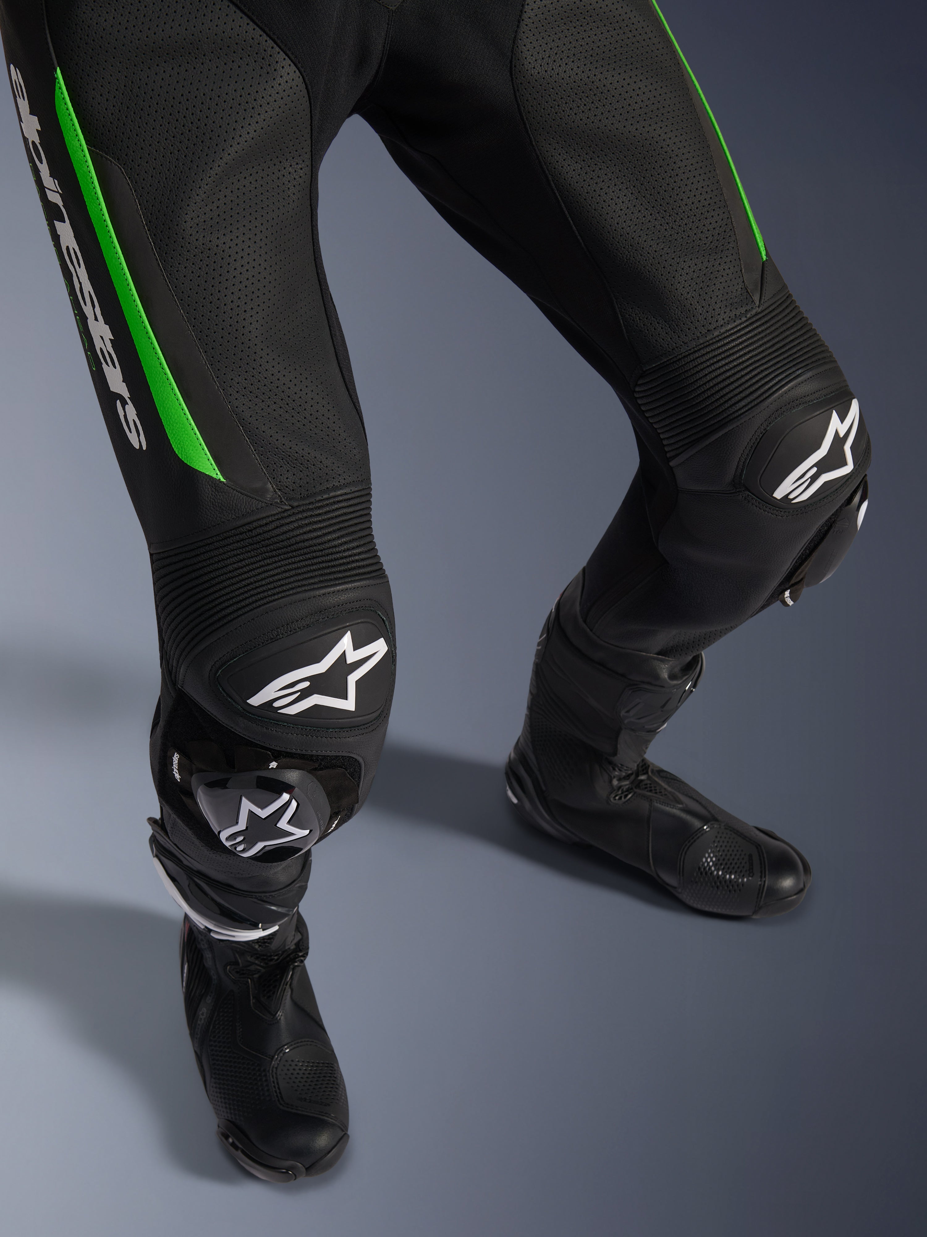 Alpinestars GP Force V2 Leather Suit - 1 Pezzo, primo piano delle gambe in nero e verde, con costruzione in pelle traforata, pannelli a fisarmonica elasticizzati sulle ginocchia, marchio Alpinestars bianco e saponette integrate, indossata con stivali da racing neri.