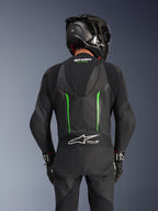 Vista posteriore di un pilota che indossa la tuta in pelle Alpinestars GP Force V2 in Black Green, con gobba aerodinamica, logo Tech-Air Ready sul pannello a fisarmonica della zona lombare e costruzione in pelle traforata, abbinata a un casco da corsa nero in fibra di carbonio.