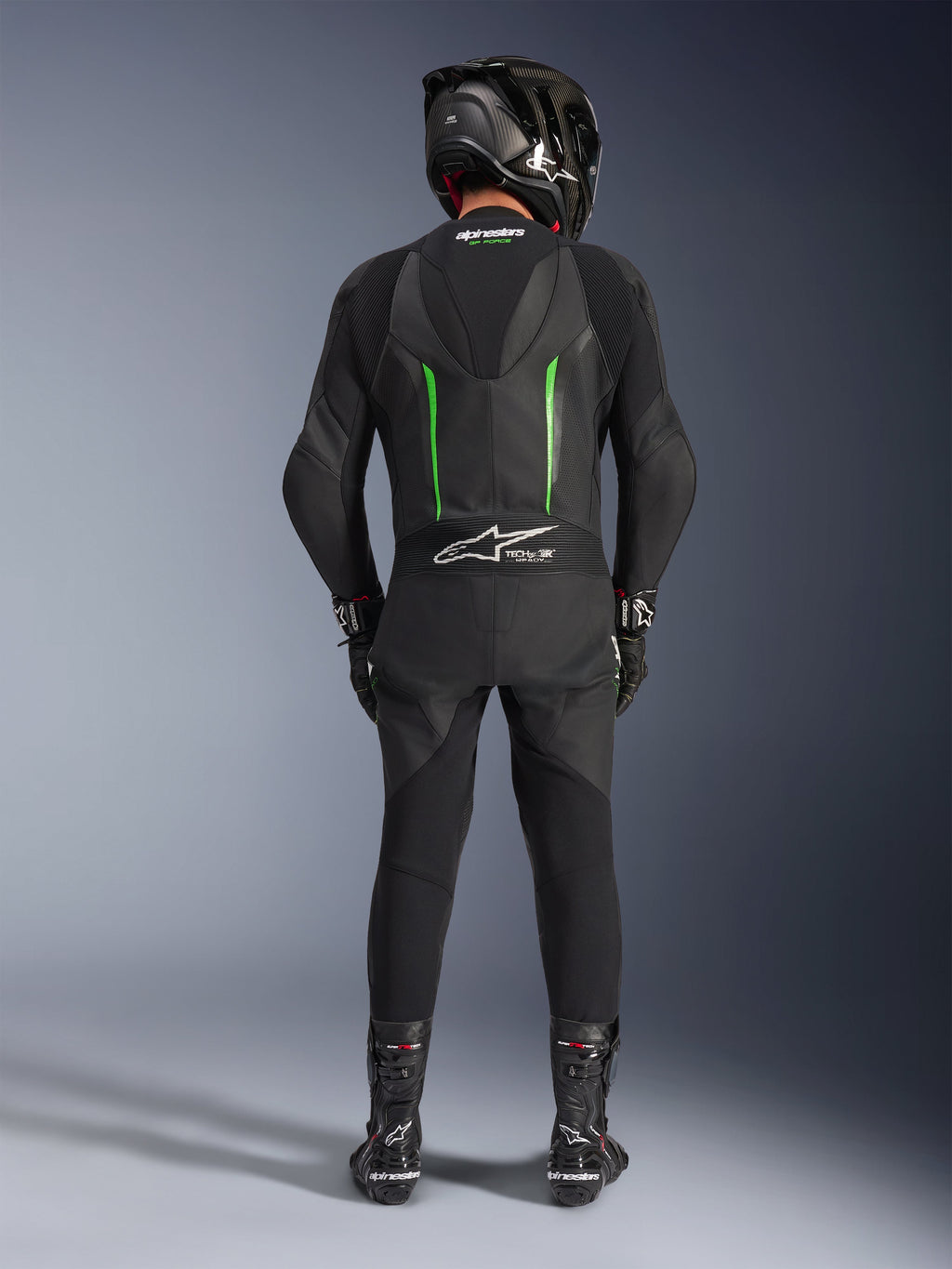 Pilota visto da dietro che indossa la tuta in pelle Alpinestars GP Force V2 - 1 pezzo in nero e verde, abbinata a casco in fibra di carbonio nero, guanti da corsa neri e stivali neri, con gobba aerodinamica e logo Tech-Air Ready.