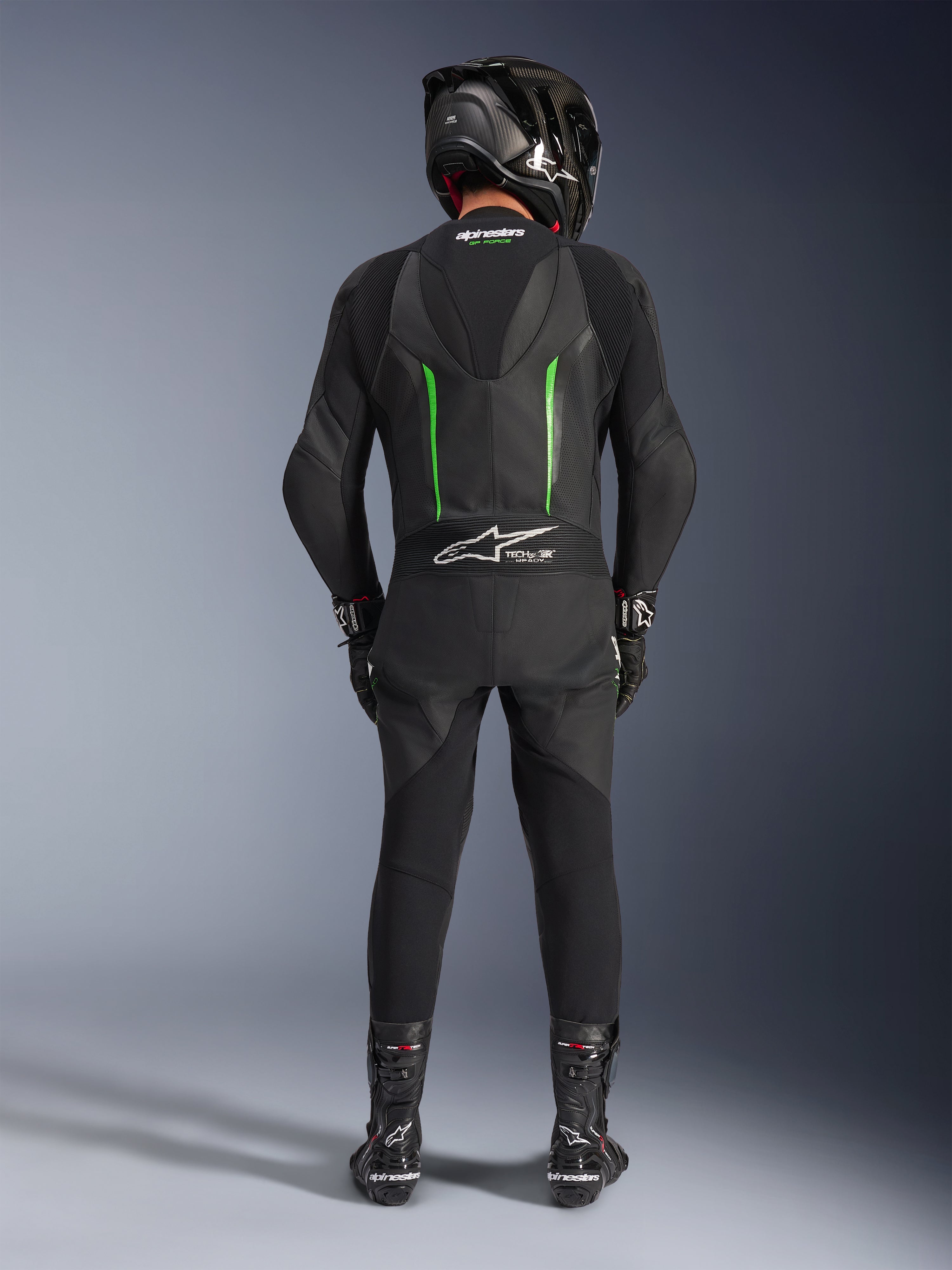 Pilota visto da dietro che indossa la tuta in pelle Alpinestars GP Force V2 - 1 pezzo in nero e verde, abbinata a casco in fibra di carbonio nero, guanti da corsa neri e stivali neri, con gobba aerodinamica e logo Tech-Air Ready.
