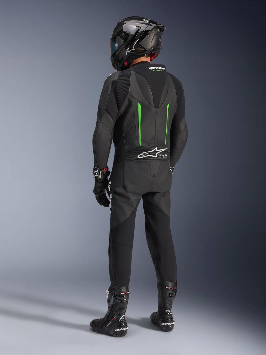 Pilota visto da dietro con tuta in pelle Alpinestars GP Force V2, 1 pezzo, nero con dettagli verdi, gobba aerodinamica, abbinata a casco nero in fibra di carbonio, stivali e guanti da corsa neri, abbigliamento per competizioni motociclistiche ad alte prestazioni