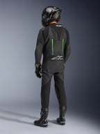 Pilota visto da dietro con tuta in pelle Alpinestars GP Force V2, 1 pezzo, nero con dettagli verdi, gobba aerodinamica, abbinata a casco nero in fibra di carbonio, stivali e guanti da corsa neri, abbigliamento per competizioni motociclistiche ad alte prestazioni