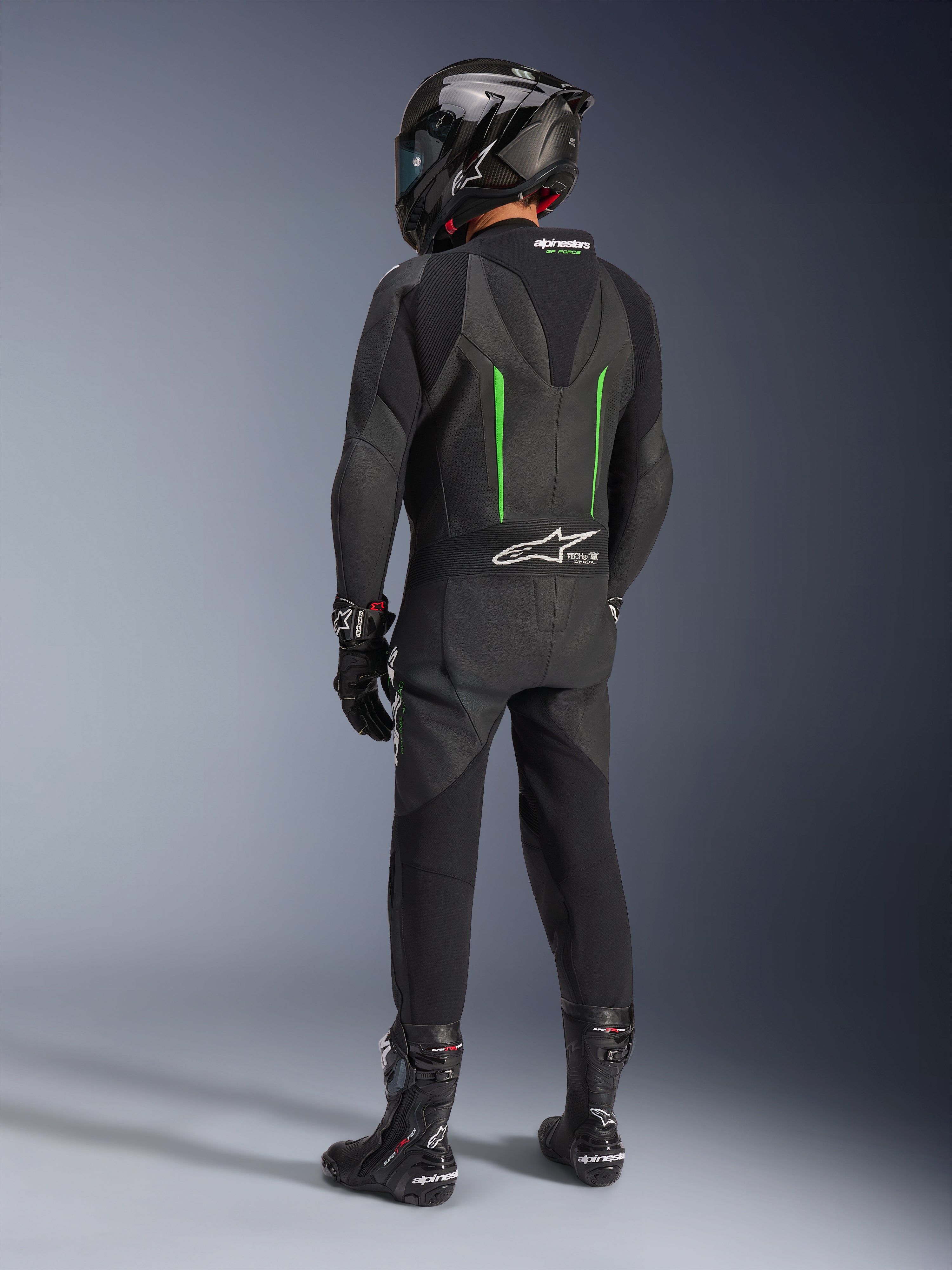 Pilota visto da dietro con tuta in pelle Alpinestars GP Force V2, 1 pezzo, nero con dettagli verdi, gobba aerodinamica, abbinata a casco nero in fibra di carbonio, stivali e guanti da corsa neri, abbigliamento per competizioni motociclistiche ad alte prestazioni