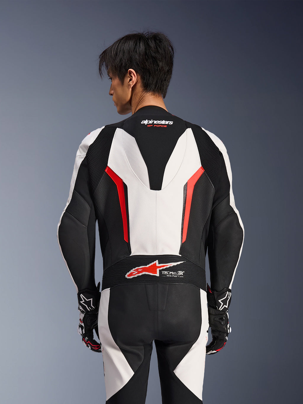 Alpinestars GP Force V2 Leather Suit 1 Pezzo, bianco, nero e rosso fluo, vista posteriore su modello maschile, con gobba aerodinamica, pannelli in pelle traforata e logo Tech-Air Ready sulla parte bassa della schiena, abbigliamento racing professionale con guanti neri e bianchi