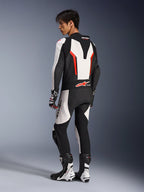 Pilota di moto che indossa la tuta in pelle Alpinestars GP Force V2 - 1 Pezzo in bianco, nero e rosso fluo, vista posteriore-laterale che mostra la gobba aerodinamica e i pannelli in pelle traforata, abbinata a stivali e guanti da corsa coordinati, progettata per la pista e la guida ad alte prestazioni.