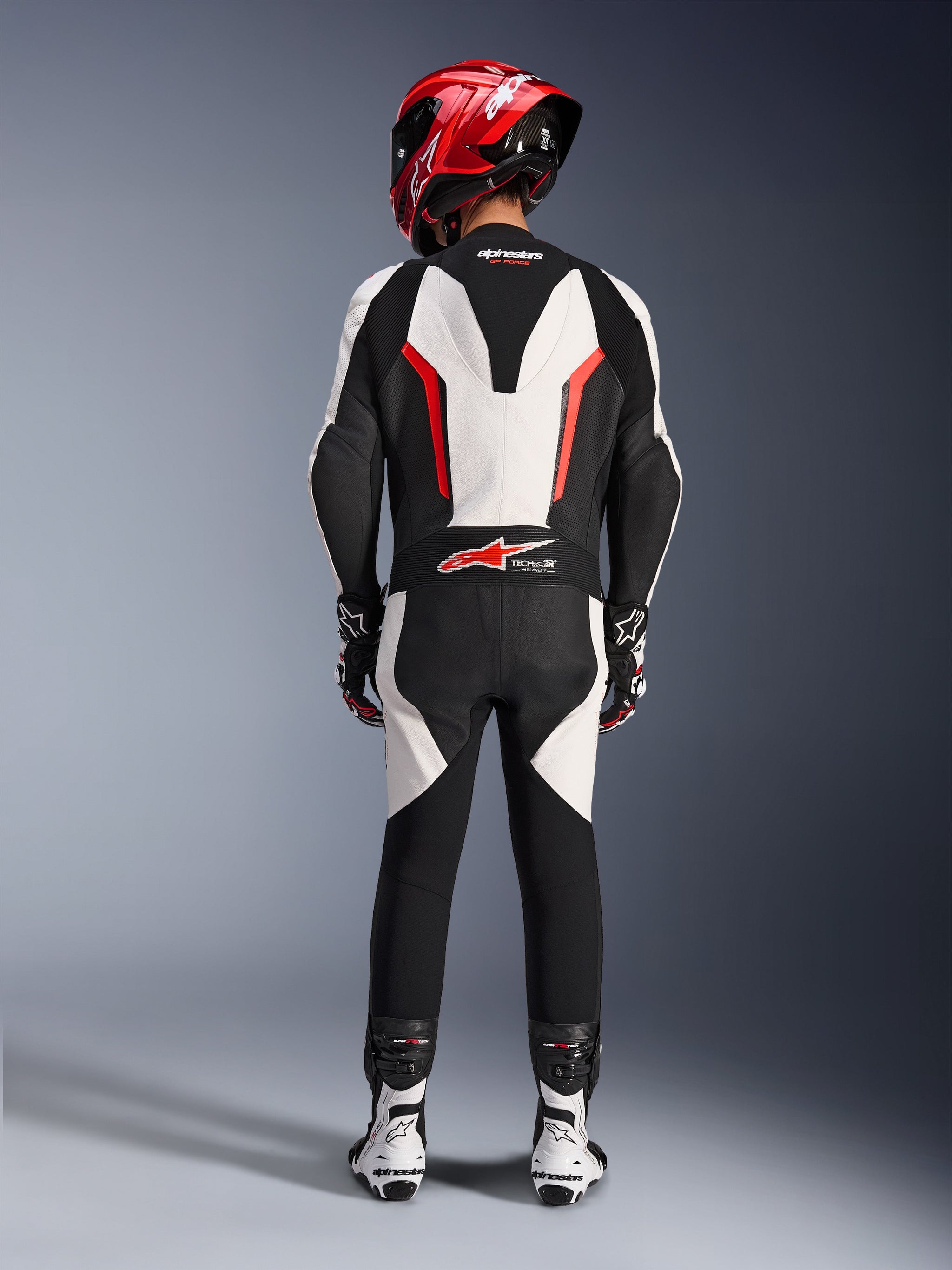 Tuta in pelle Alpinestars GP Force V2 - Monopezzo, Bianco Nero Rosso Fluo, vista posteriore che evidenzia la gobba aerodinamica e il marchio Tech-Air Ready, indossata con casco da corsa rosso e stivali bianchi e neri, tuta in pelle per prestazioni in pista.