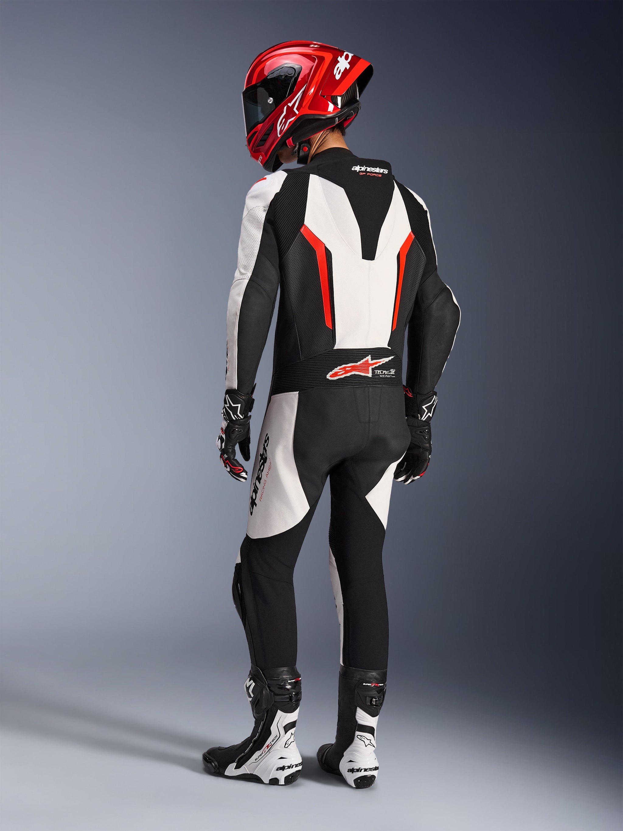 Alpinestars GP Force V2 Leather Suit - Monopezzo, Bianco Nero Rosso Fluo, vista posteriore del pilota con equipaggiamento da gara completo, inclusi casco rosso, stivali e guanti bianchi e neri, evidenziando la gobba aerodinamica e i pannelli elasticizzati ergonomici.