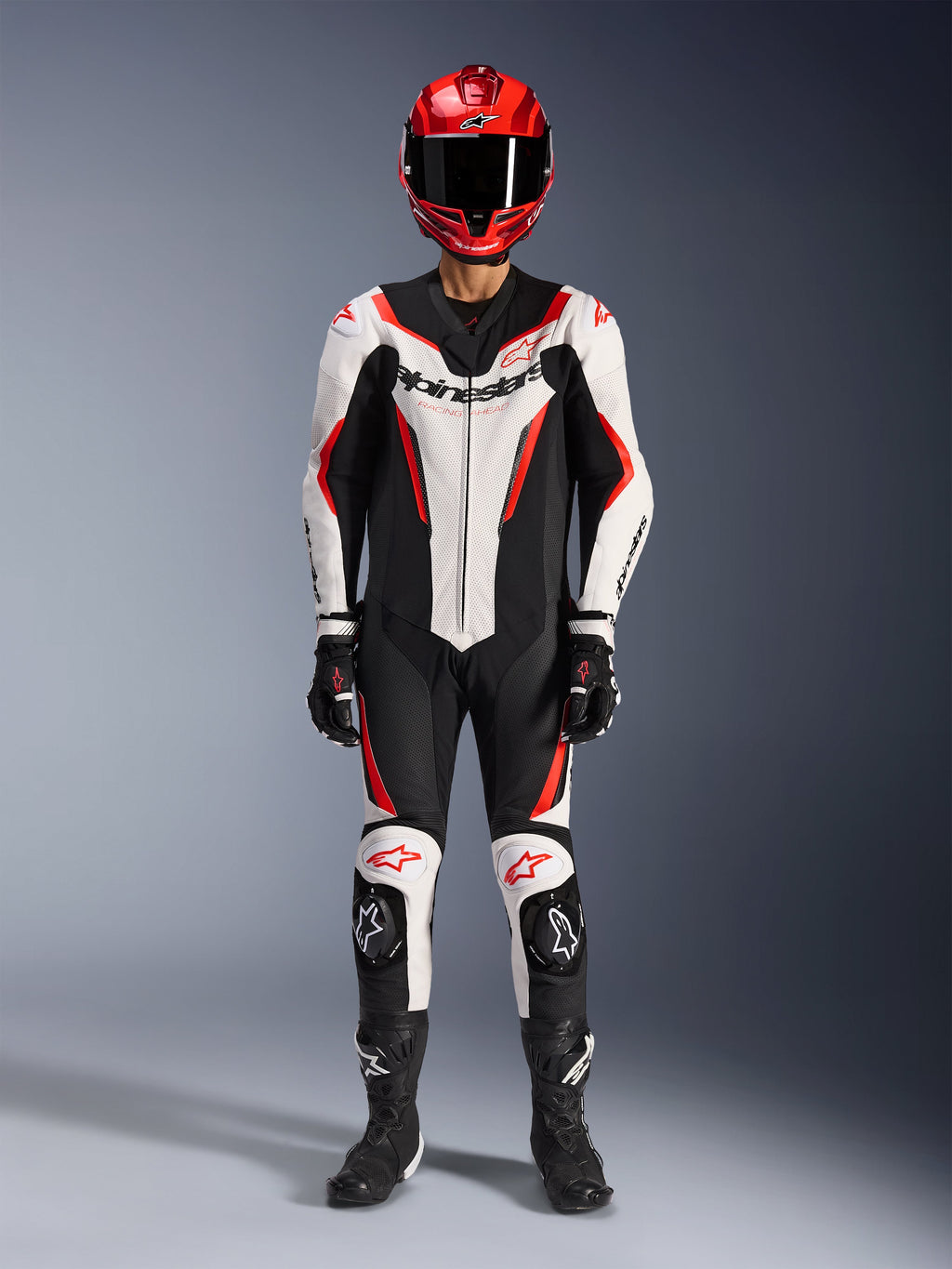 Tuta in pelle Alpinestars GP Force V2 monopezzo, bianco, nero e rosso fluo, equipaggiamento racing completo con casco rosso, guanti e stivali neri, caratterizzata da design aerodinamico e loghi Alpinestars su petto e ginocchia