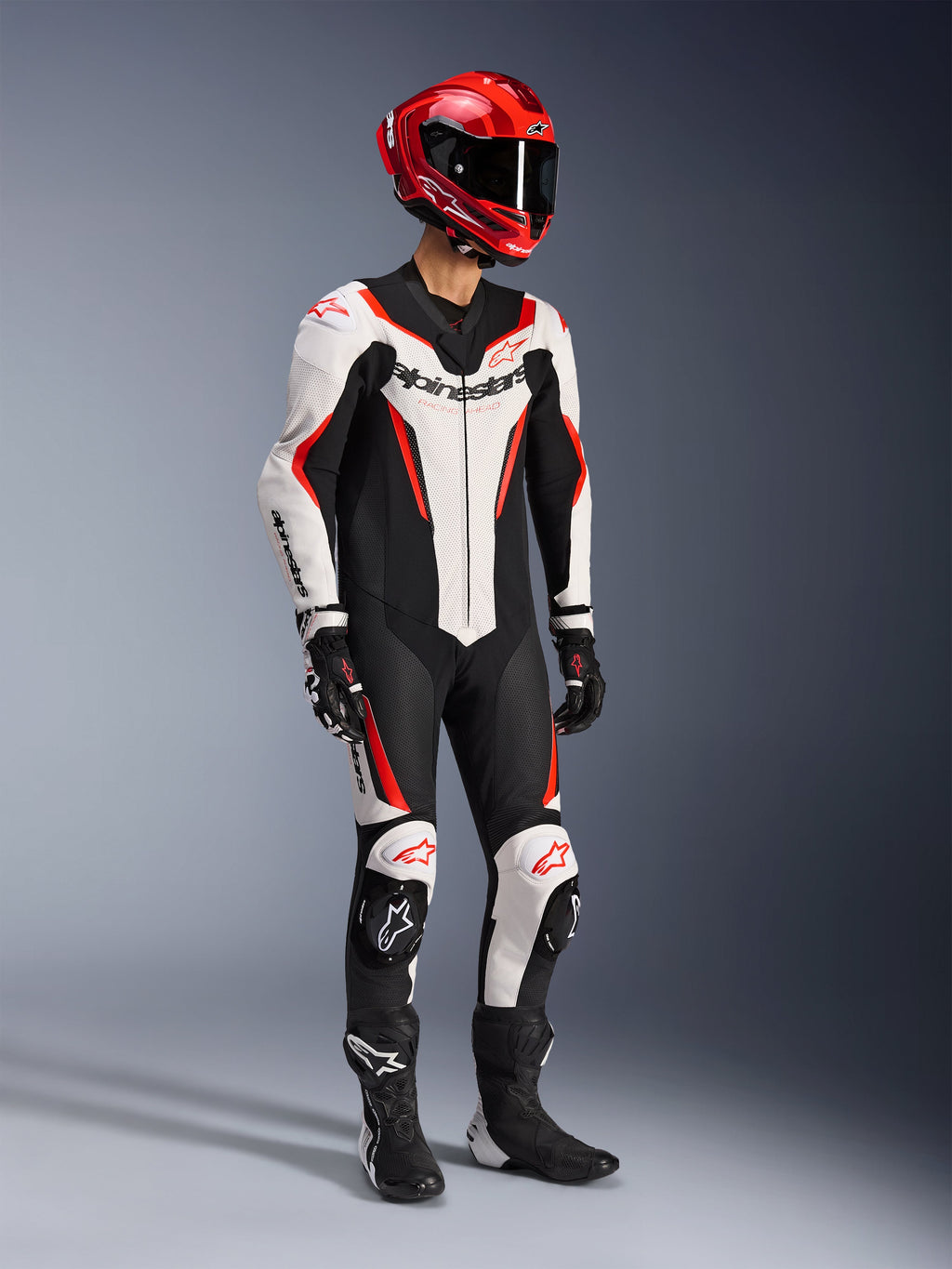 Alpinestars GP Force V2 Leather Suit 1-Piece in bianco, nero e rosso fluo, scatto in studio a figura intera di un pilota con equipaggiamento completo da gara, comprensivo di casco rosso, guanti neri e stivali neri, costruzione aerodinamica in pelle bovina con loghi e slider per ginocchia integrati.