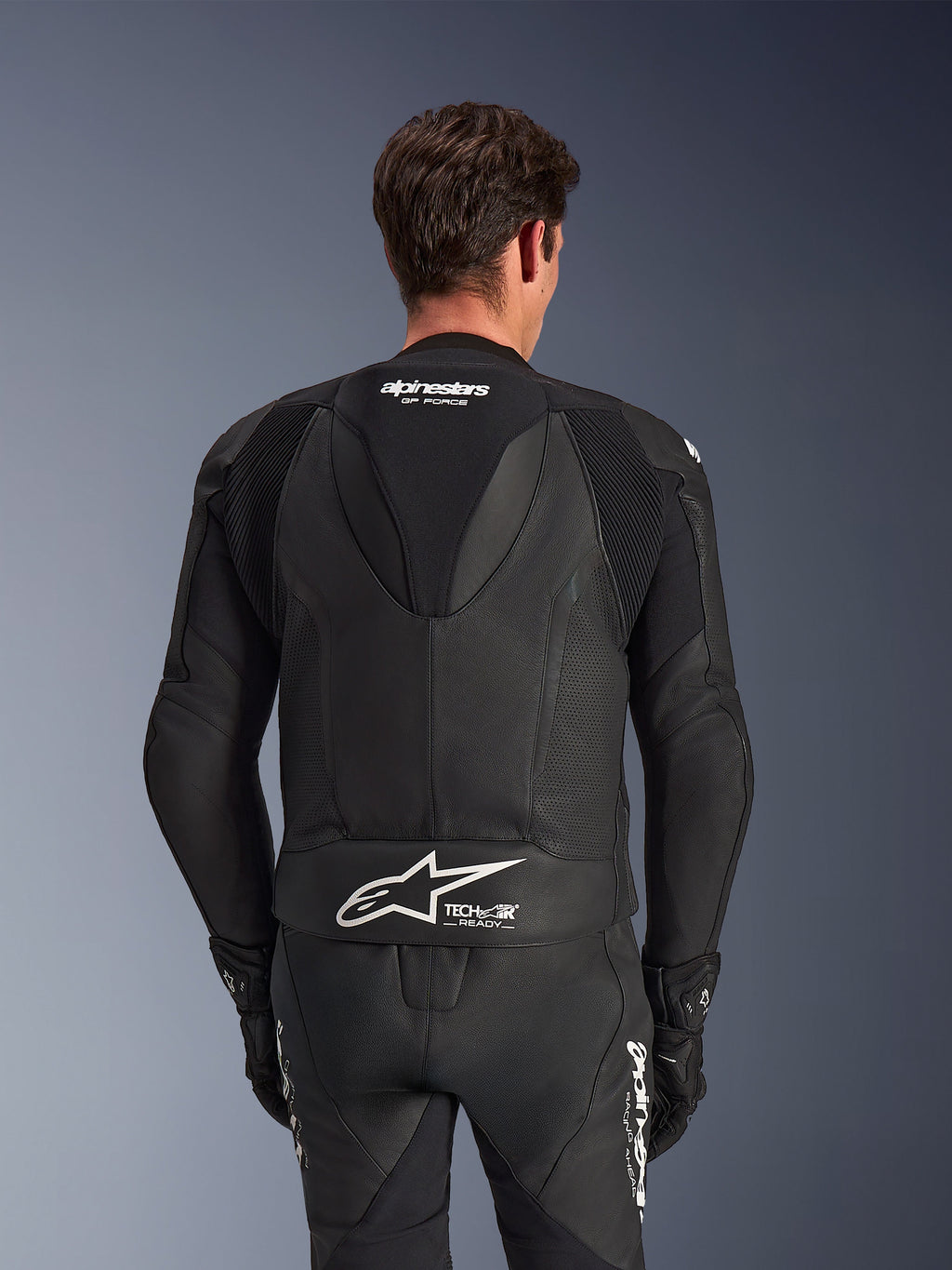 Tuta in pelle Alpinestars GP Force V2 2 Pezzi, nera, vista posteriore del pilota, con pannelli in pelle traforata, gobba aerodinamica con marchio GP Force e logo Alpinestars bianco con scritta Tech-Air Ready sulla parte bassa della schiena