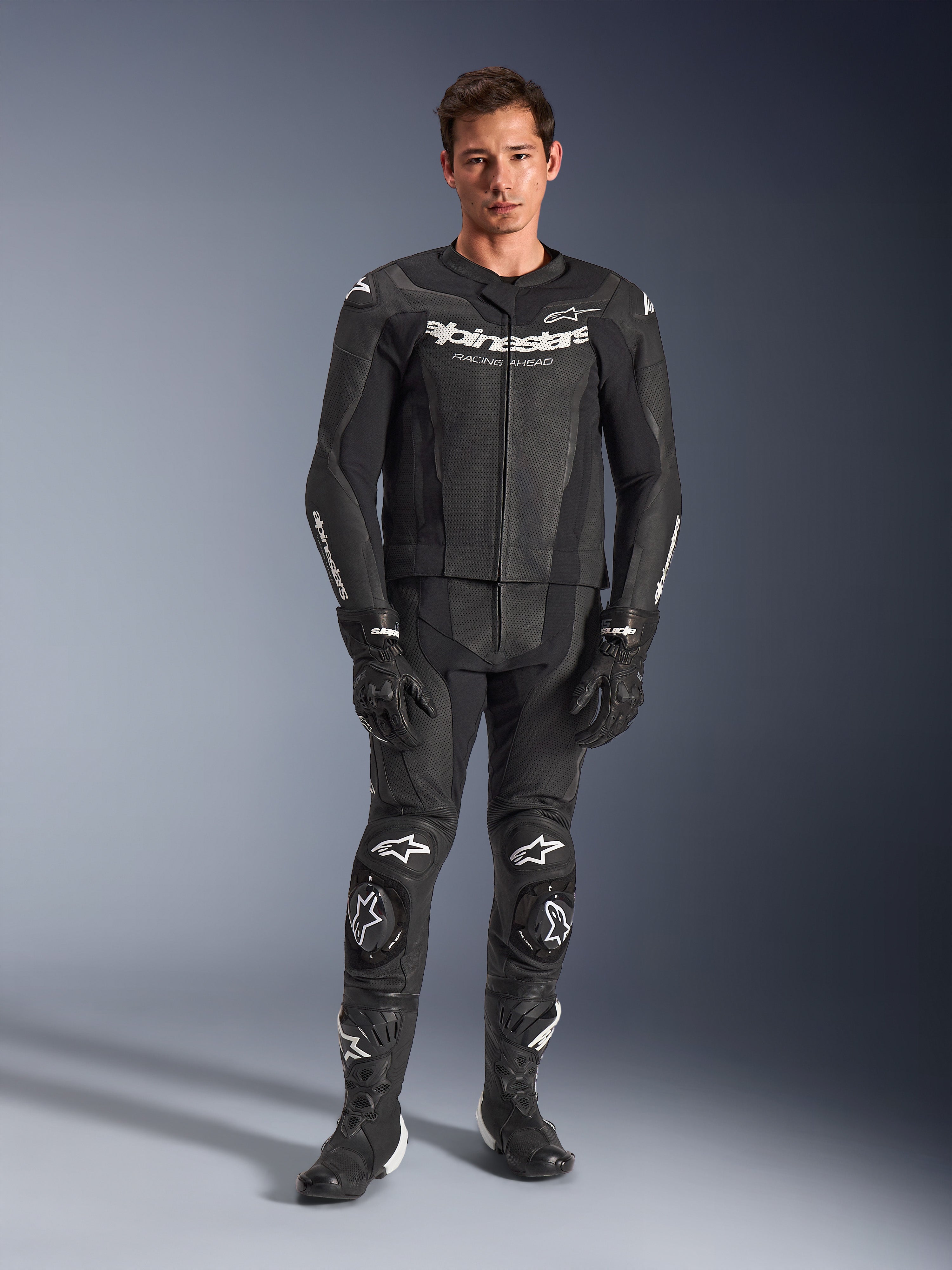 Alpinestars GP Force V2, tuta in pelle a 2 pezzi, nero con loghi bianchi, tuta integrale da corsa, struttura in pelle premium con slider per ginocchia rinforzati e pannelli traforati, indossata con guanti e stivali neri, vista a figura intera in studio