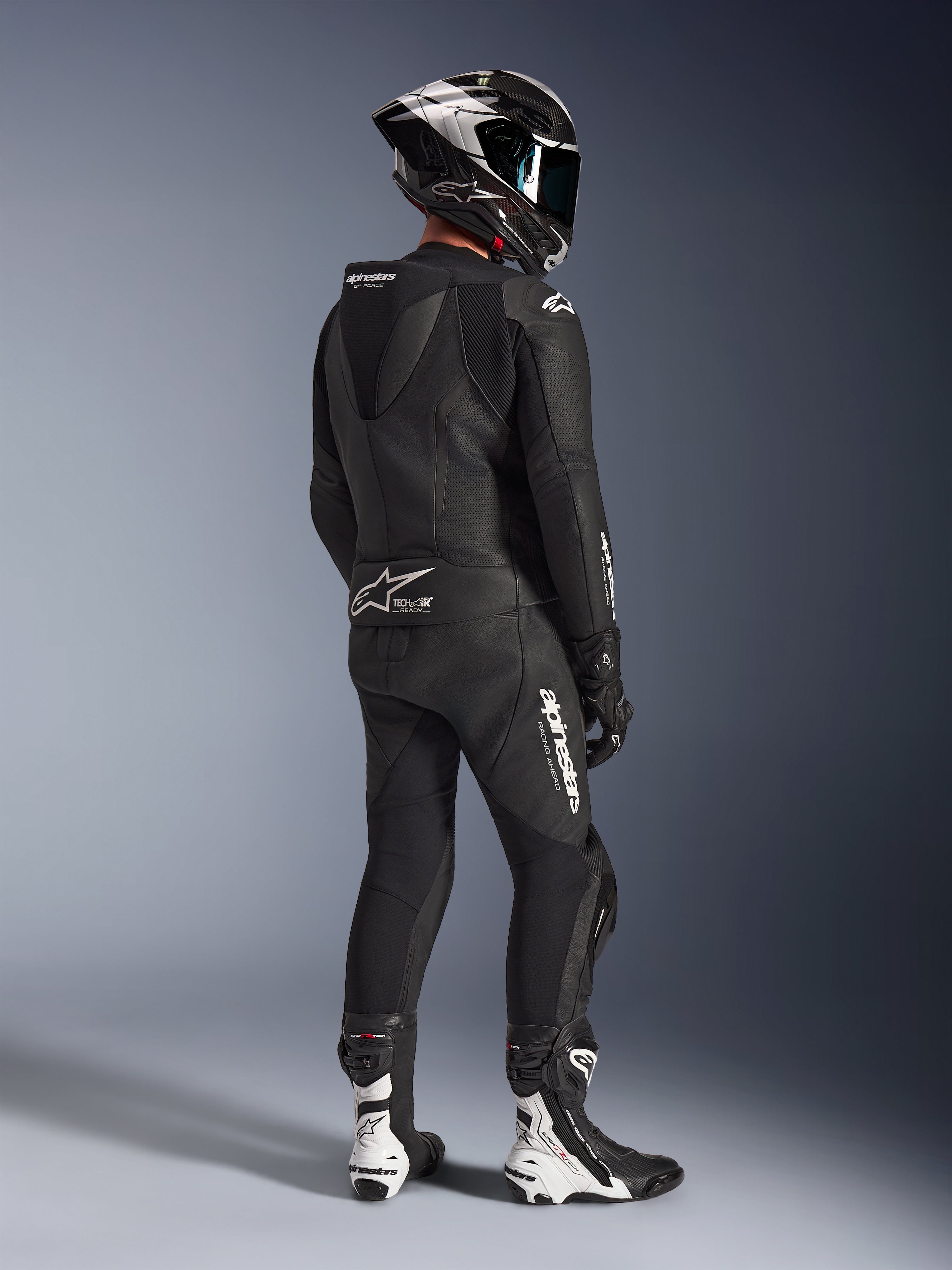Alpinestars GP Force V2 2-Piece Leather Suit, tuta in pelle a due pezzi, nera, vista posteriore di un pilota con attrezzatura da corsa completa, gobba aerodinamica, loghi bianchi e pelle traforata, abbinata a casco in fibra di carbonio e stivali racing neri e bianchi