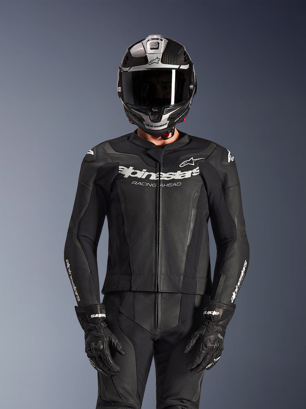 Pilota che indossa la tuta in pelle Alpinestars GP Force V2 2 pezzi in nero, costruzione in pelle traforata con logo bianco 'Racing Ahead' sul petto, casco integrale argento e fibra di carbonio, guanti neri, abbigliamento professionale per guida sportiva e in pista