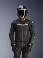 Pilota che indossa la tuta in pelle Alpinestars GP Force V2 2 pezzi in nero, costruzione in pelle traforata con logo bianco 'Racing Ahead' sul petto, casco integrale argento e fibra di carbonio, guanti neri, abbigliamento professionale per guida sportiva e in pista