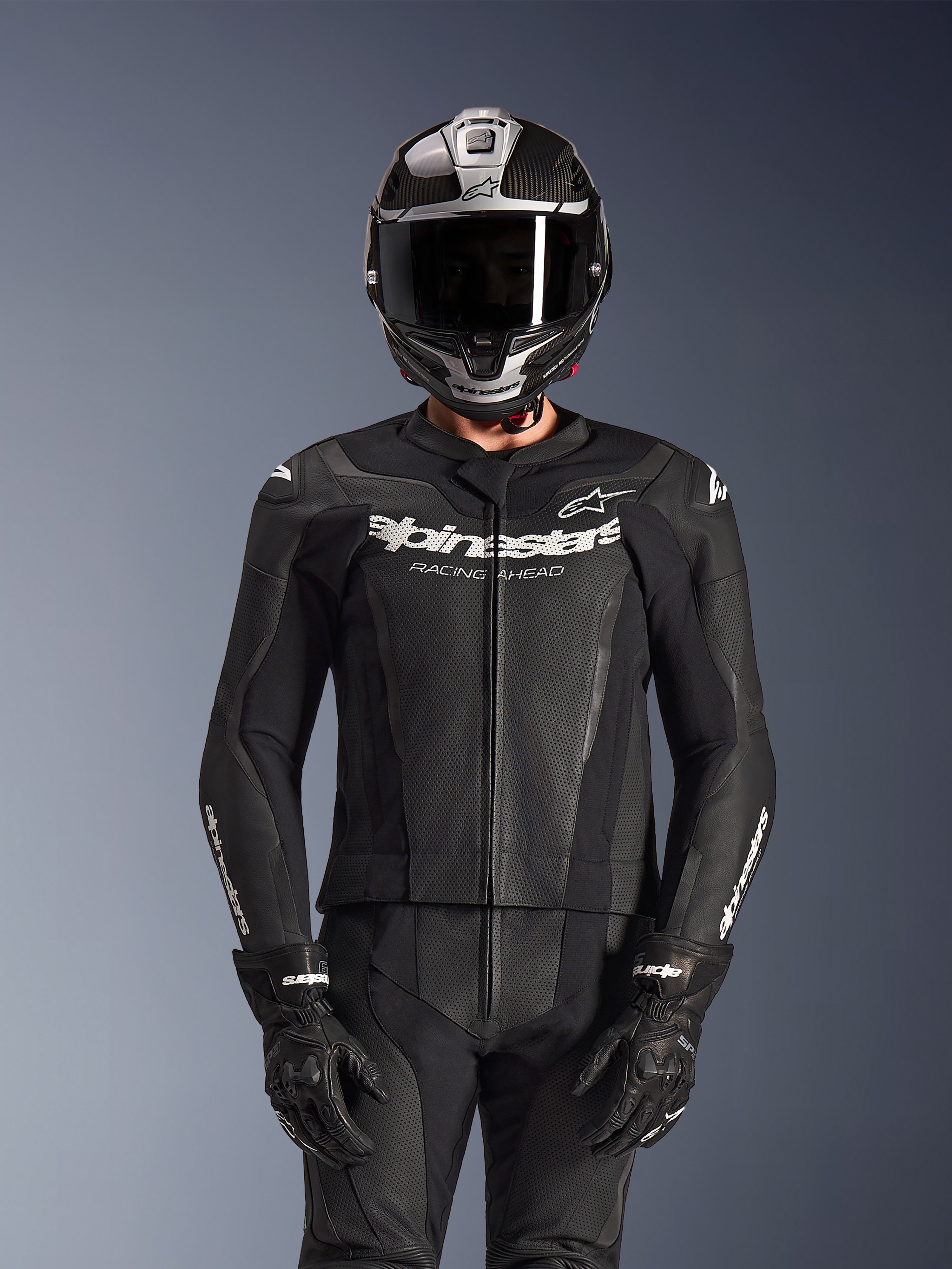 Pilota che indossa la tuta in pelle Alpinestars GP Force V2 2 pezzi in nero, costruzione in pelle traforata con logo bianco 'Racing Ahead' sul petto, casco integrale argento e fibra di carbonio, guanti neri, abbigliamento professionale per guida sportiva e in pista