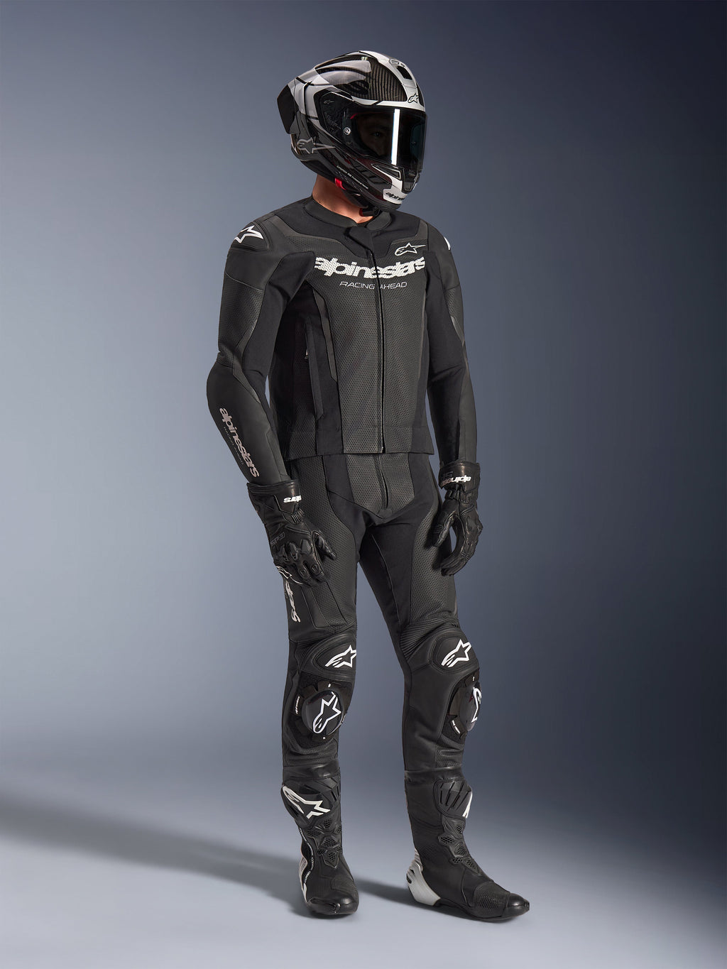 Alpinestars GP Force V2 2-Piece Leather Suit, tuta da moto in pelle nera, indossata con casco integrale, guanti e stivali, caratterizzata da pelle traforata, gobba aerodinamica e loghi Alpinestars bianchi su petto, braccia e ginocchia.