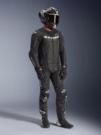 Alpinestars GP Force V2 2-Piece Leather Suit, tuta da moto in pelle nera, indossata con casco integrale, guanti e stivali, caratterizzata da pelle traforata, gobba aerodinamica e loghi Alpinestars bianchi su petto, braccia e ginocchia.
