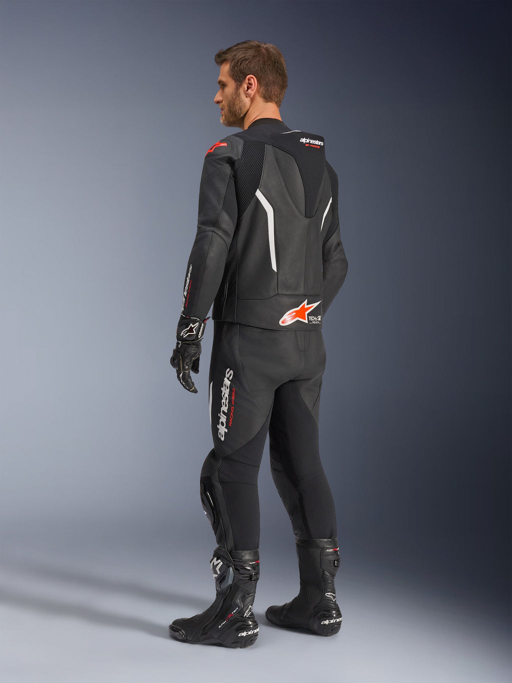Tuta in pelle Alpinestars GP Force V2 2 pezzi, nera con dettagli bianco e rosso fluo, vista posteriore di tre quarti su modello, con gobba aerodinamica, pannelli in pelle traforata e logo Alpinestars in evidenza, indossata con stivali e guanti da corsa neri.