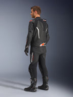 Tuta in pelle Alpinestars GP Force V2 2 pezzi, nera con dettagli bianco e rosso fluo, vista posteriore di tre quarti su modello, con gobba aerodinamica, pannelli in pelle traforata e logo Alpinestars in evidenza, indossata con stivali e guanti da corsa neri.