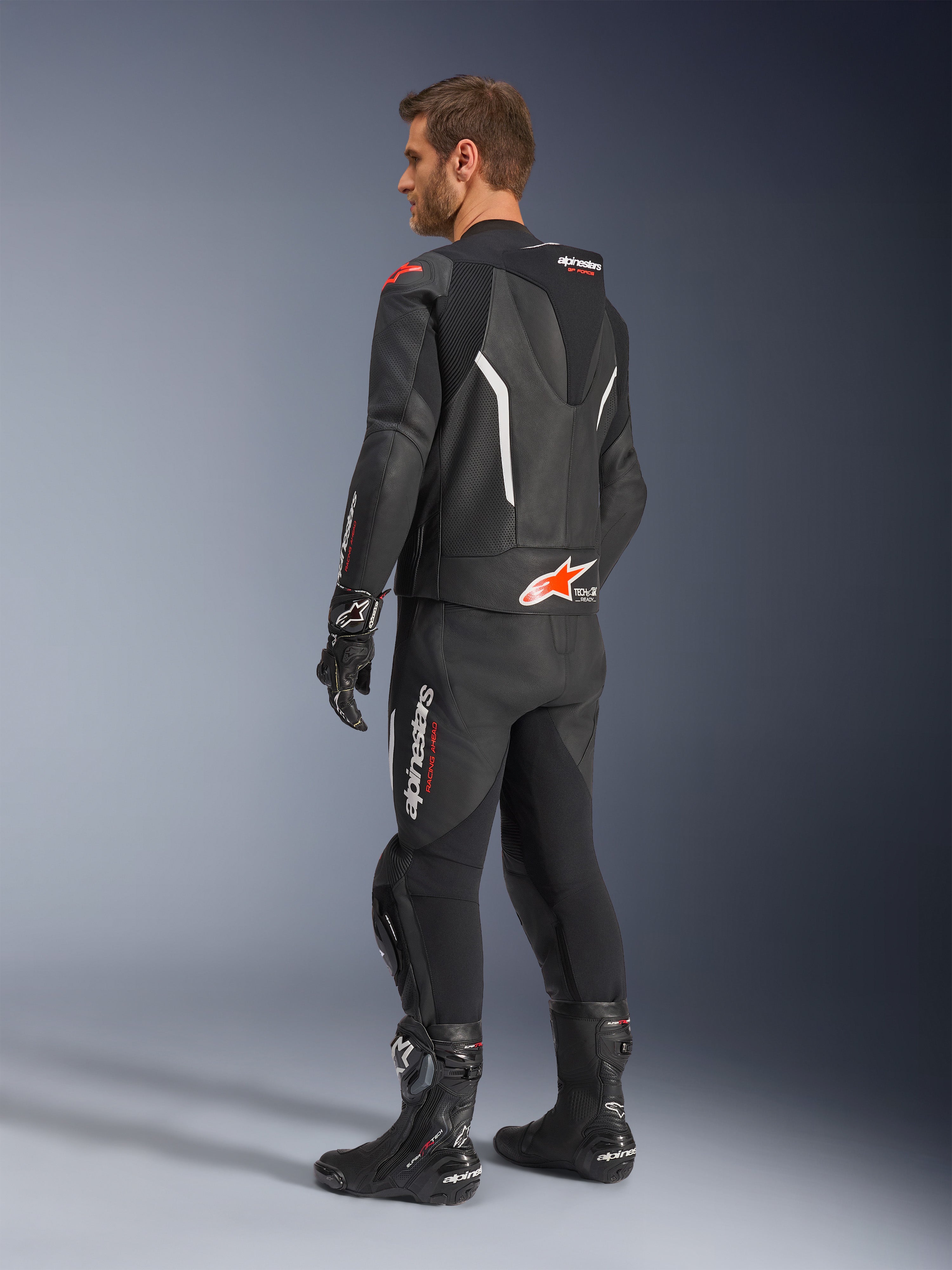 Tuta in pelle Alpinestars GP Force V2 2 pezzi, nera con dettagli bianco e rosso fluo, vista posteriore di tre quarti su modello, con gobba aerodinamica, pannelli in pelle traforata e logo Alpinestars in evidenza, indossata con stivali e guanti da corsa neri.