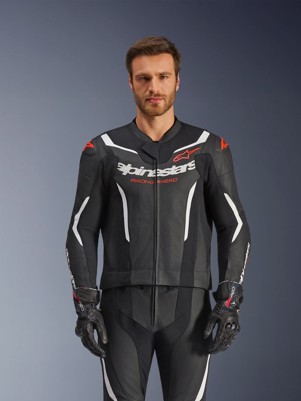 Tuta in pelle Alpinestars GP Force V2 2 pezzi, nero, bianco e rosso fluo, vista frontale di un modello che indossa la tuta in pelle traforata con guanti neri coordinati, con accenti bianchi angolari e logo Alpinestars sul petto