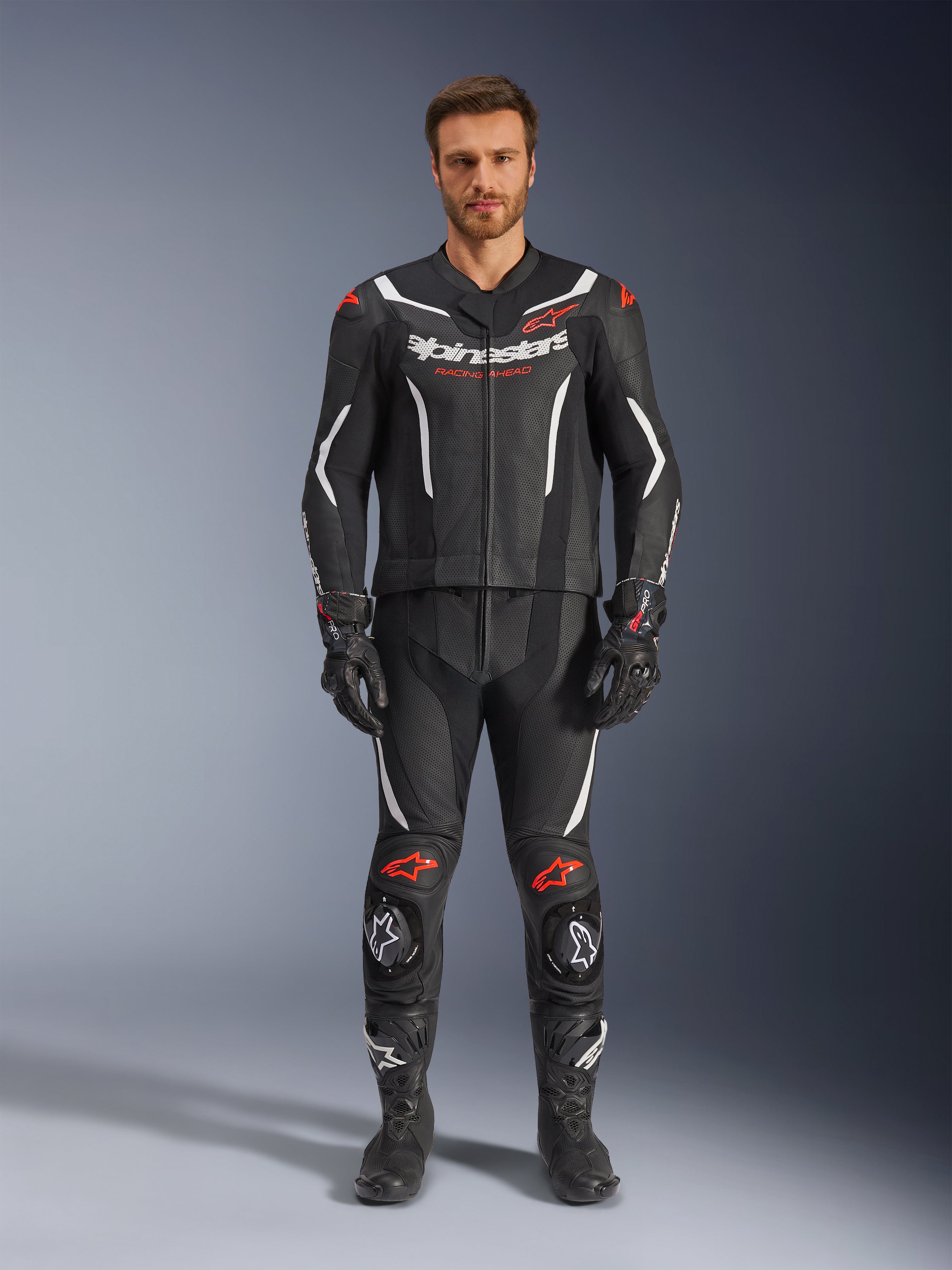 Tuta in pelle Alpinestars GP Force V2 2 pezzi, tuta da moto racing, nero, bianco e rosso fluo, indossata da un modello con guanti e stivali neri coordinati, con pannelli in pelle traforata, accenti bianco e rosso fluo e design aerodinamico per pista e guida sportiva